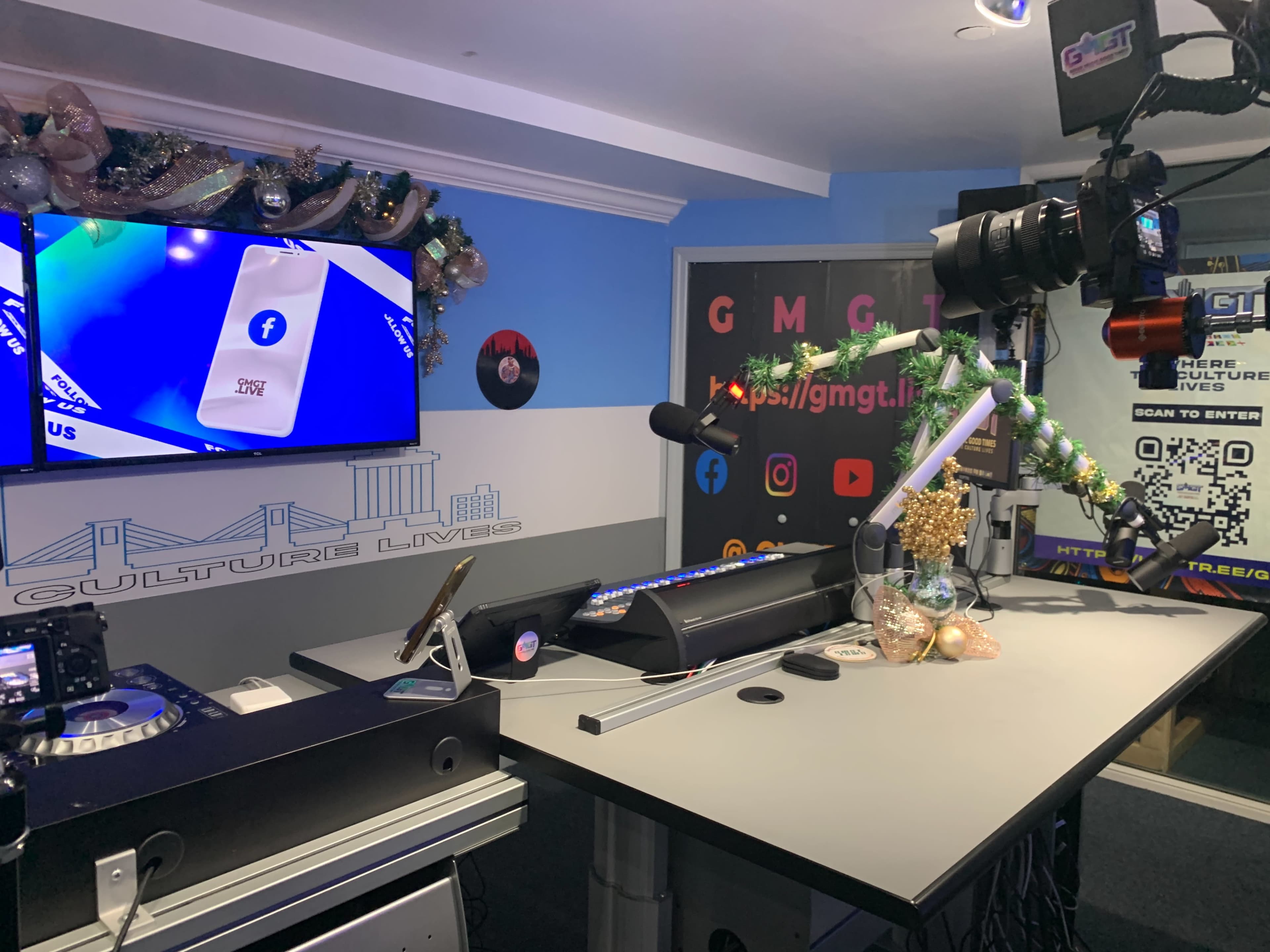 The image shows a modern broadcasting studio with a camera positioned above a desk, a television screen displaying social media content, and various decorations including a vinyl record and holiday elements.