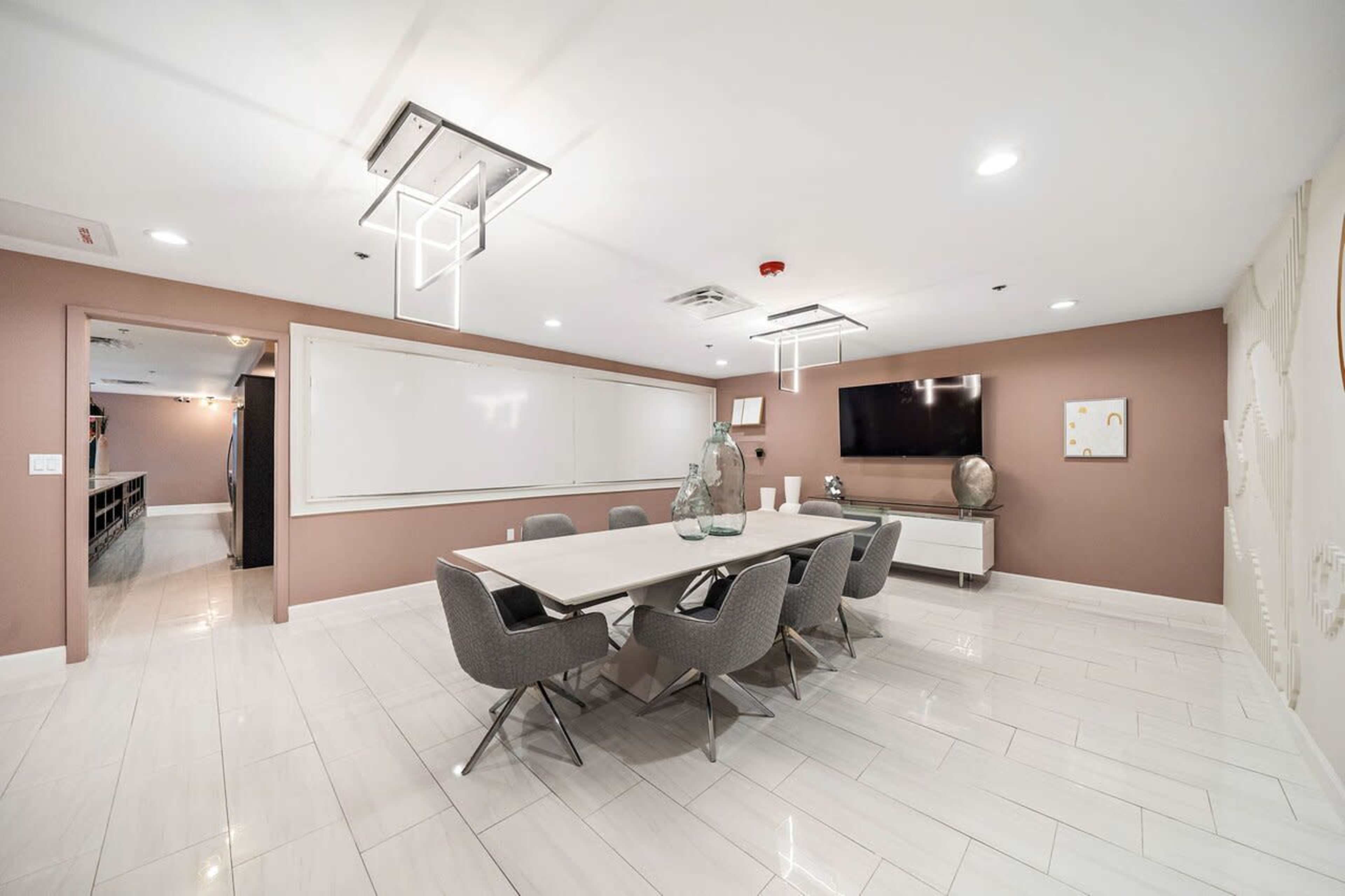A modern conference room with a long table surrounded by gray chairs, a wall-mounted TV, and a whiteboard.