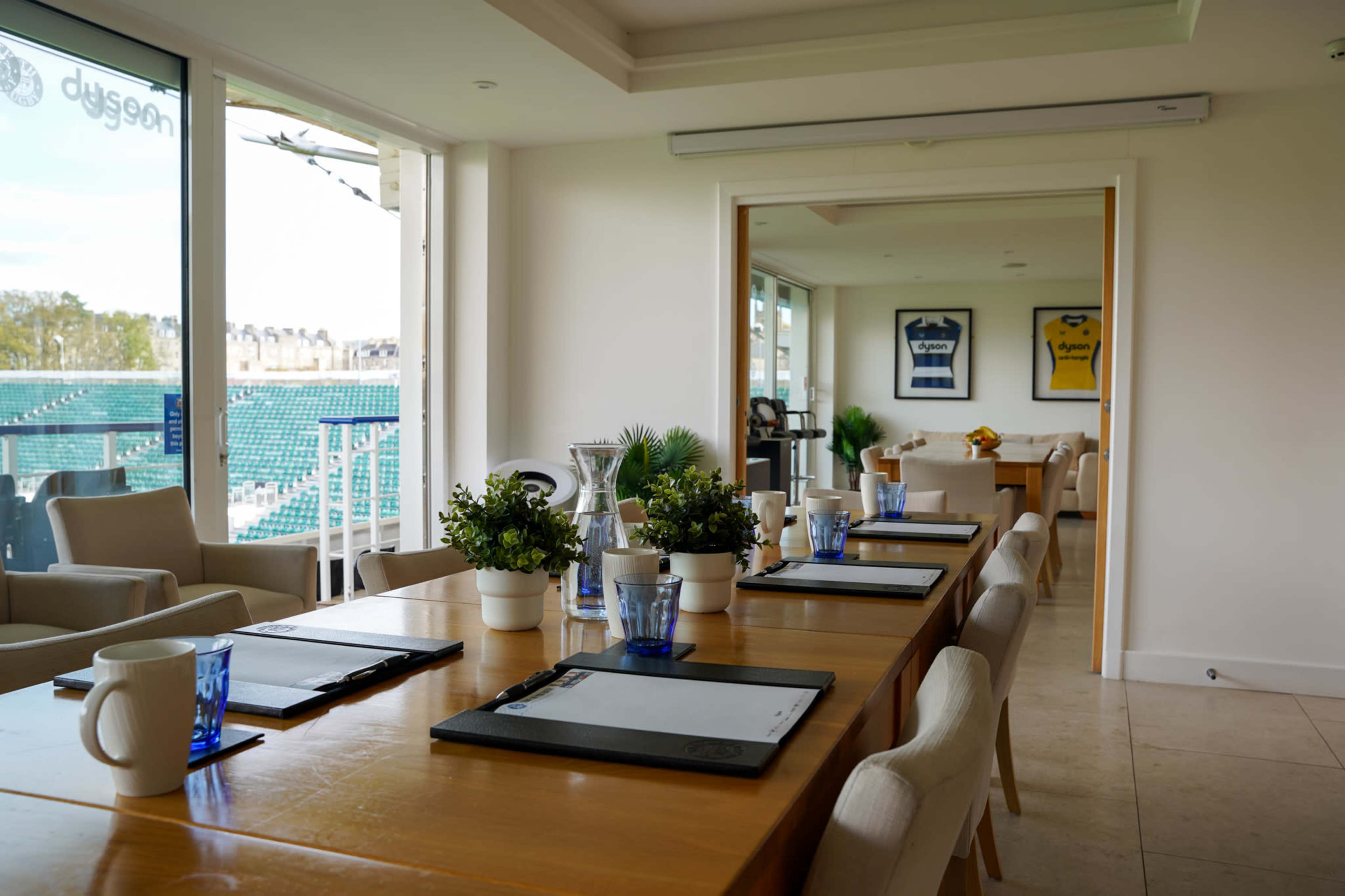 A spacious meeting room features a long wooden table set with notepads and glasses, surrounded by potted plants and large windows overlooking a sports field.