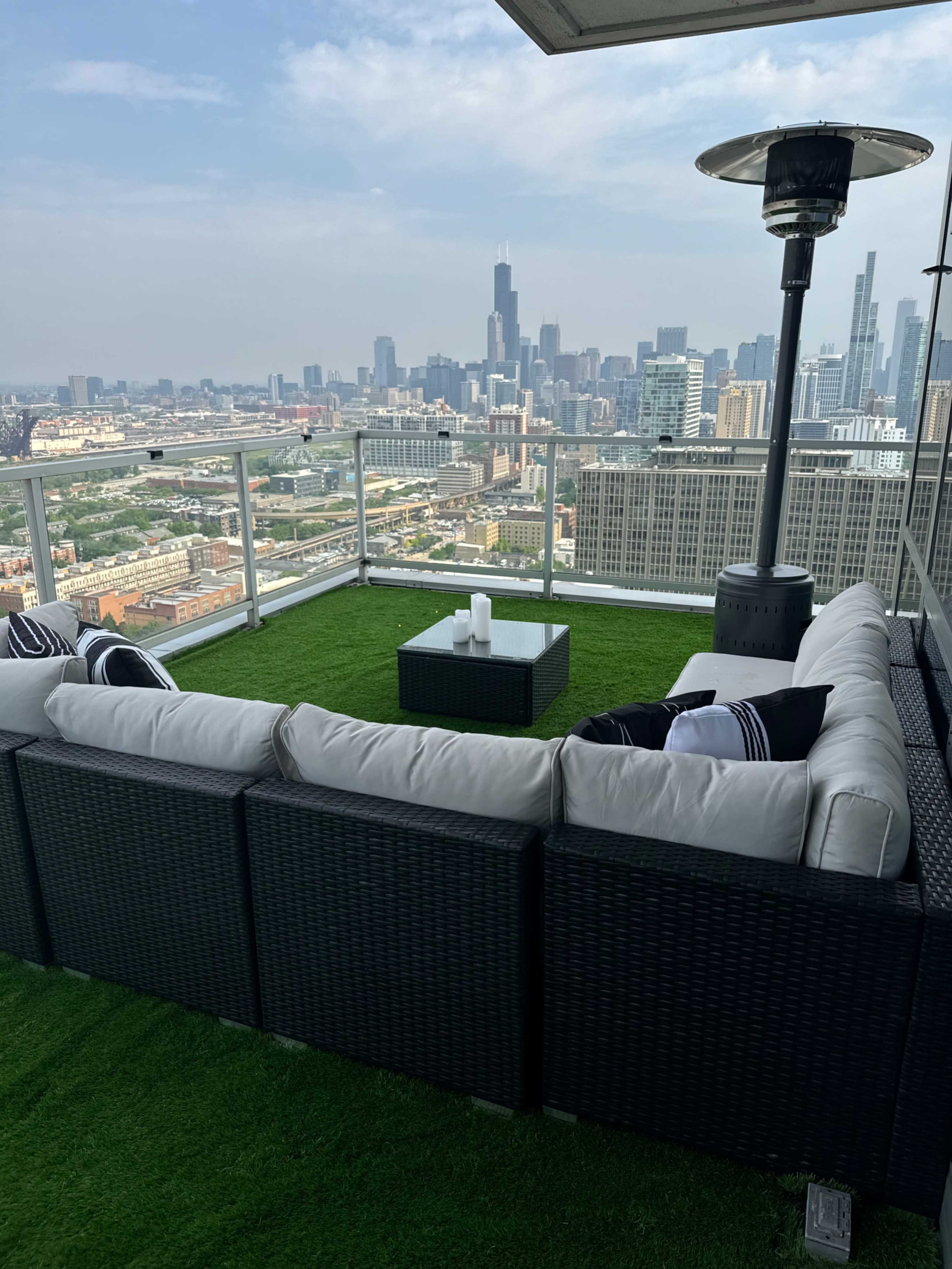 A rooftop terrace features a sectional seating area and a square coffee table on artificial grass, overlooking a city skyline.