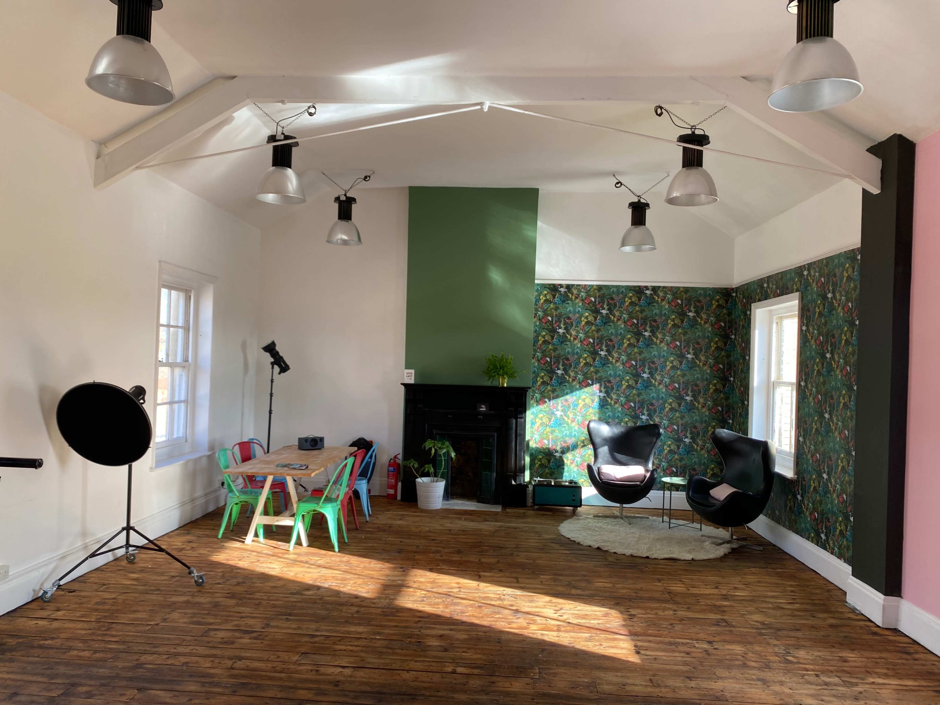 A spacious room with wooden floors, featuring a green accent wall, floral wallpaper, a black fireplace, colorful chairs around a table, and modern light fixtures.