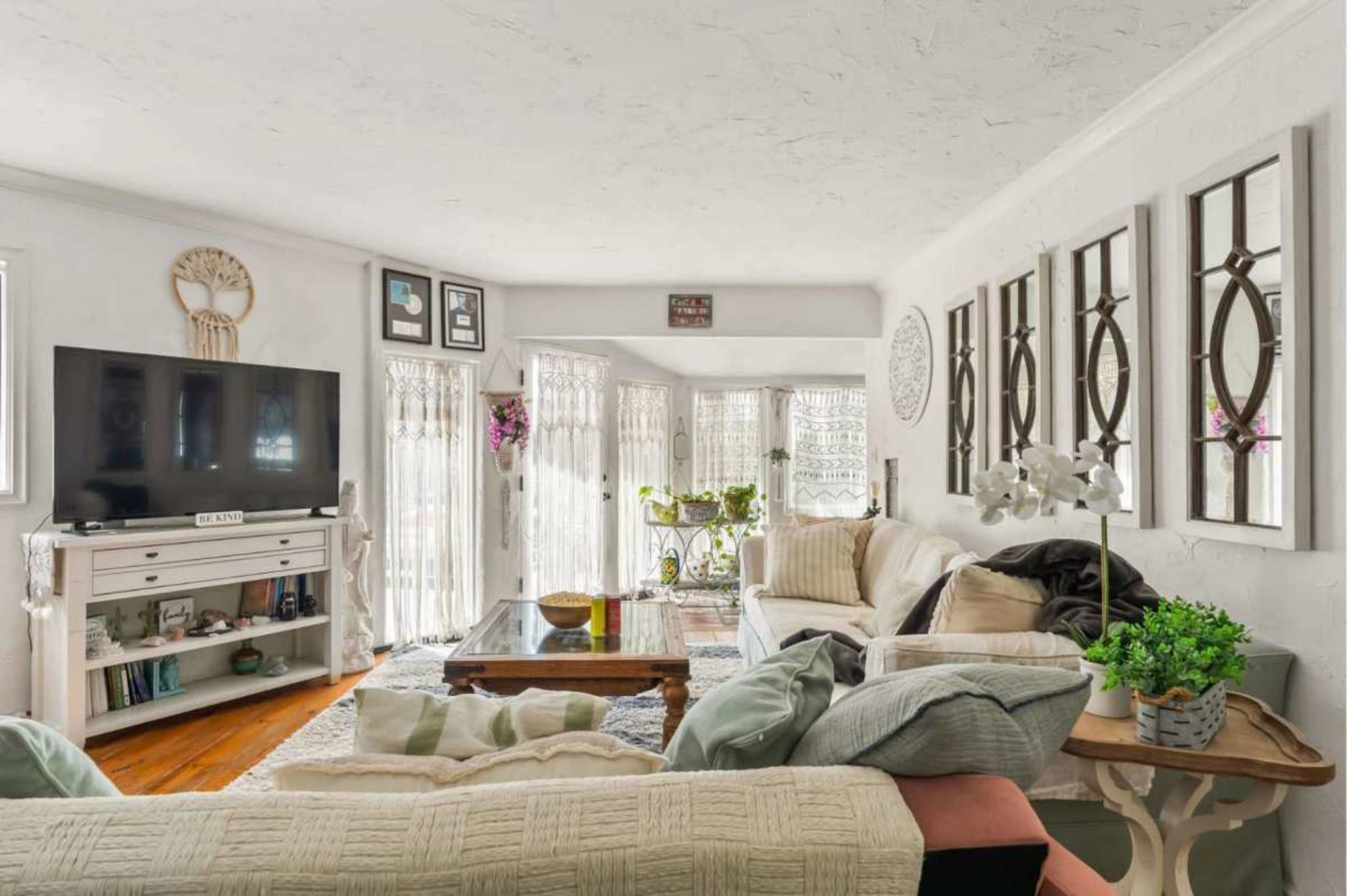 The living room features a large television on a white console, a comfortable seating area with various cushions, and windows adorned with sheer curtains that allow natural light to flow in.