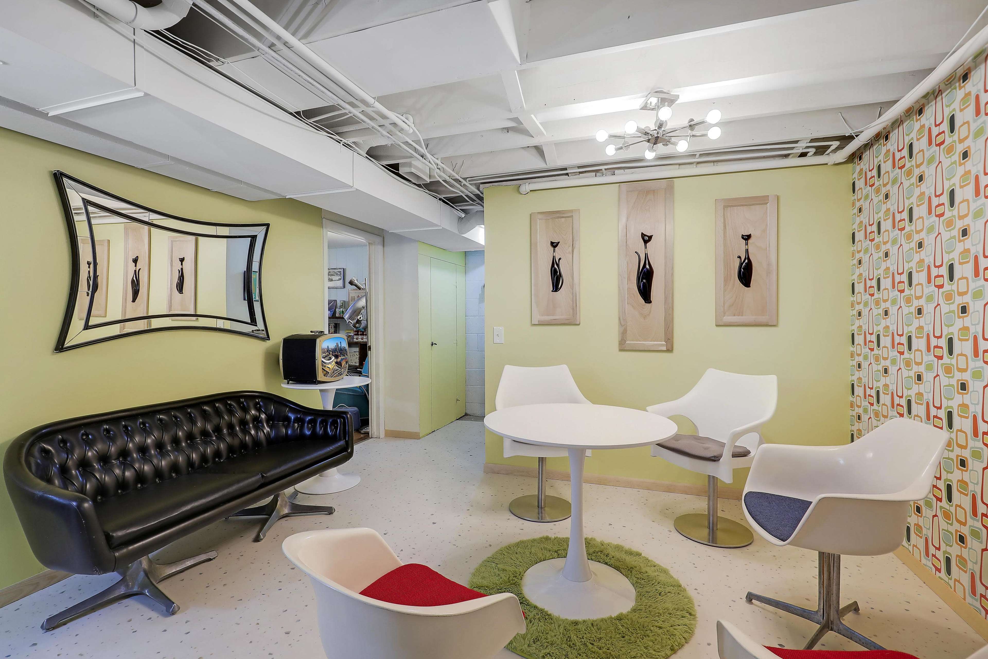 The room features a modern seating arrangement with a black tufted sofa, a round white table, and several contemporary chairs, set against a green wall with decorative art.
