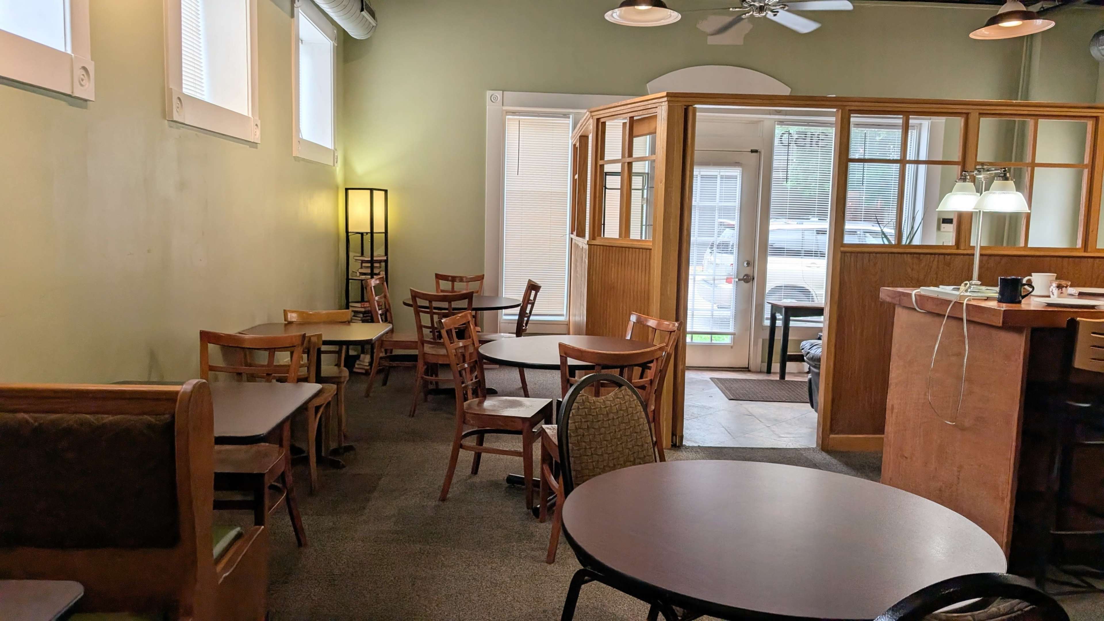 The image shows a quiet café interior featuring wooden tables and chairs arranged around a small reception area, with a doorway leading to another room.