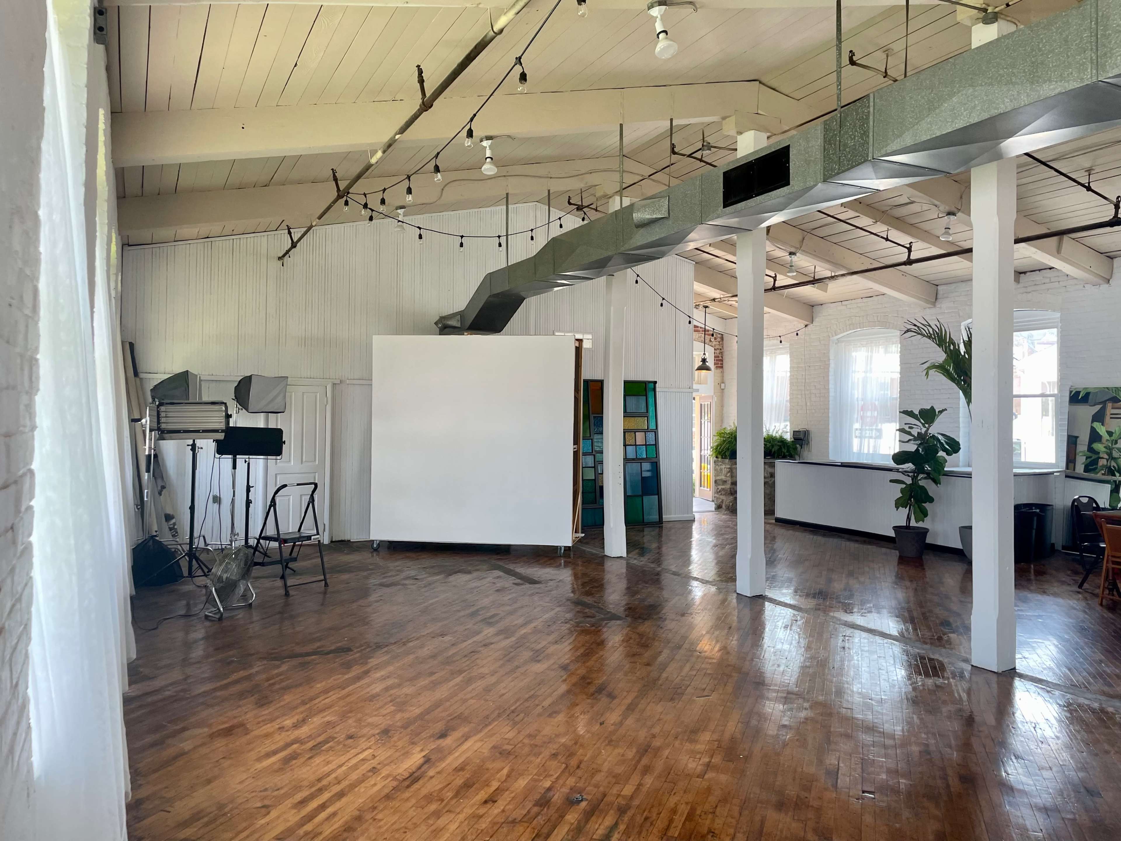 A spacious, well-lit studio with wooden floors, white walls, and large windows, featuring a central blank wall and various studio equipment.