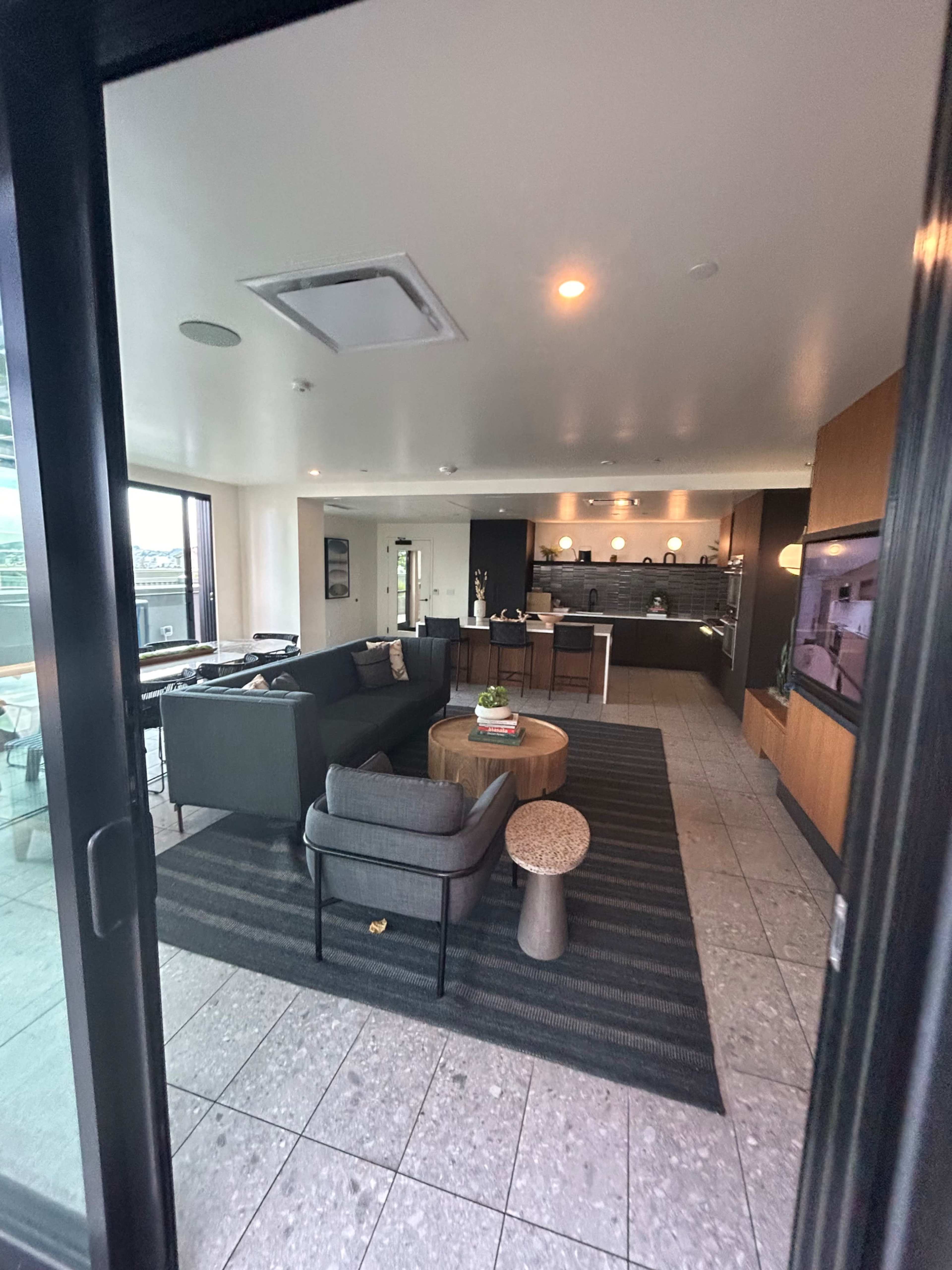 The image shows a modern, open-concept living area with a kitchen, featuring a gray couch, a round wooden coffee table, and large windows.