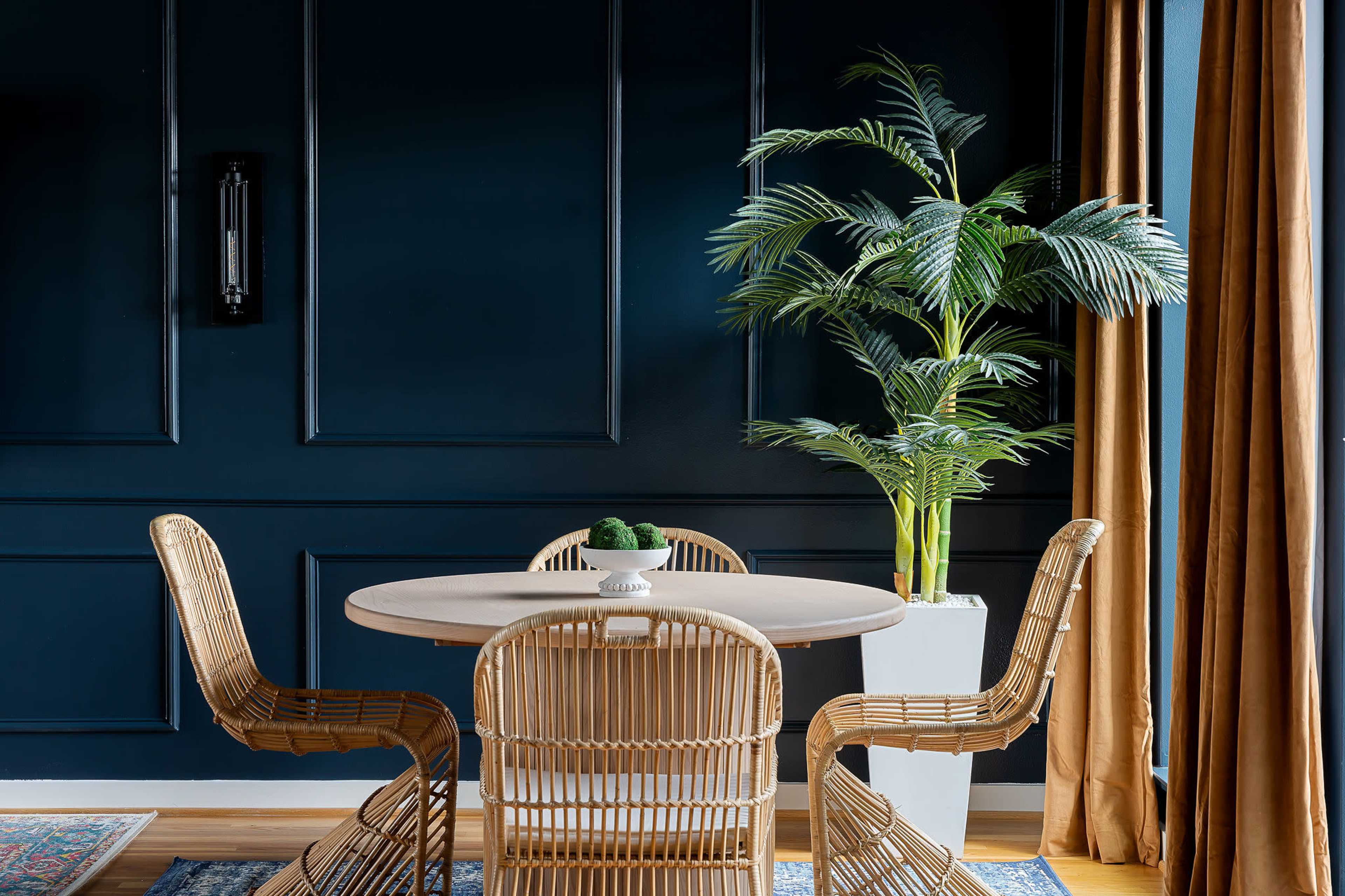 A round table with three woven chairs is set against a dark blue wall, accompanied by a tall potted plant and golden curtains.