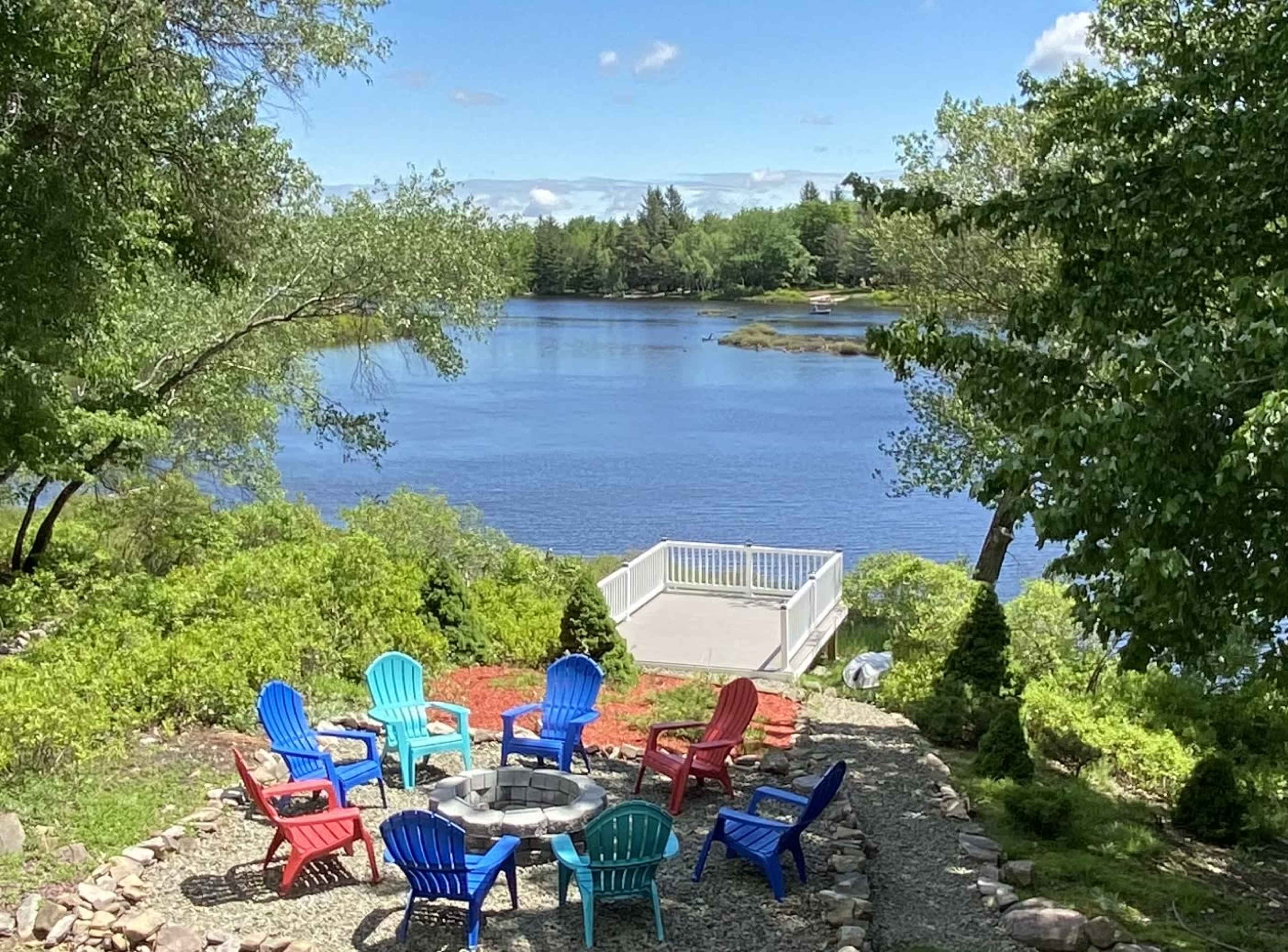 A circular arrangement of colorful chairs surrounds a stone fire pit near a calm river, with green trees and a blue sky in the background.