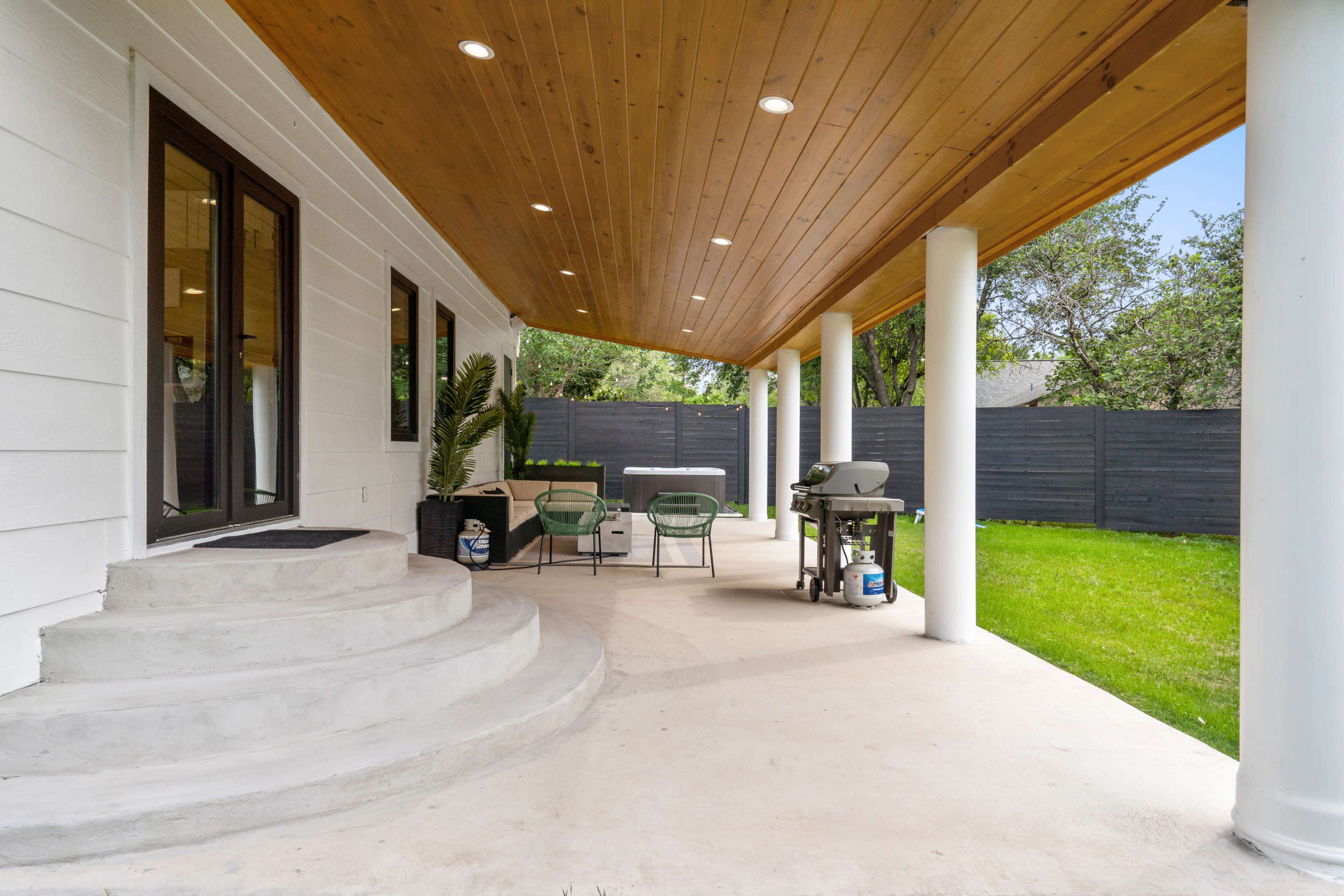A covered outdoor patio with a table, chairs, and a grill, adjacent to a grassy area and surrounded by wooden fencing.