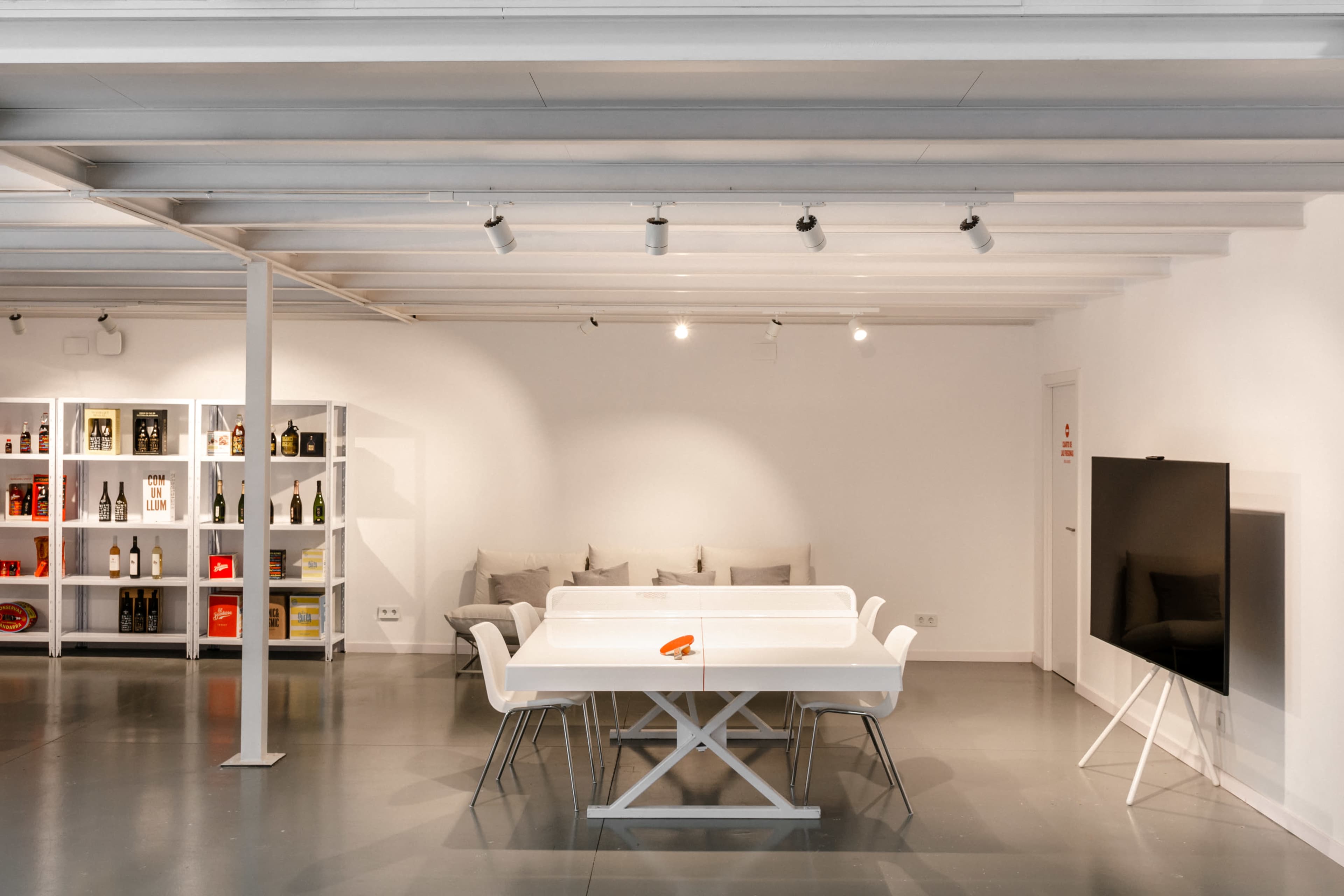 A modern meeting room with a white table and chairs, a wall-mounted TV, and shelves displaying bottles in a minimalist design.