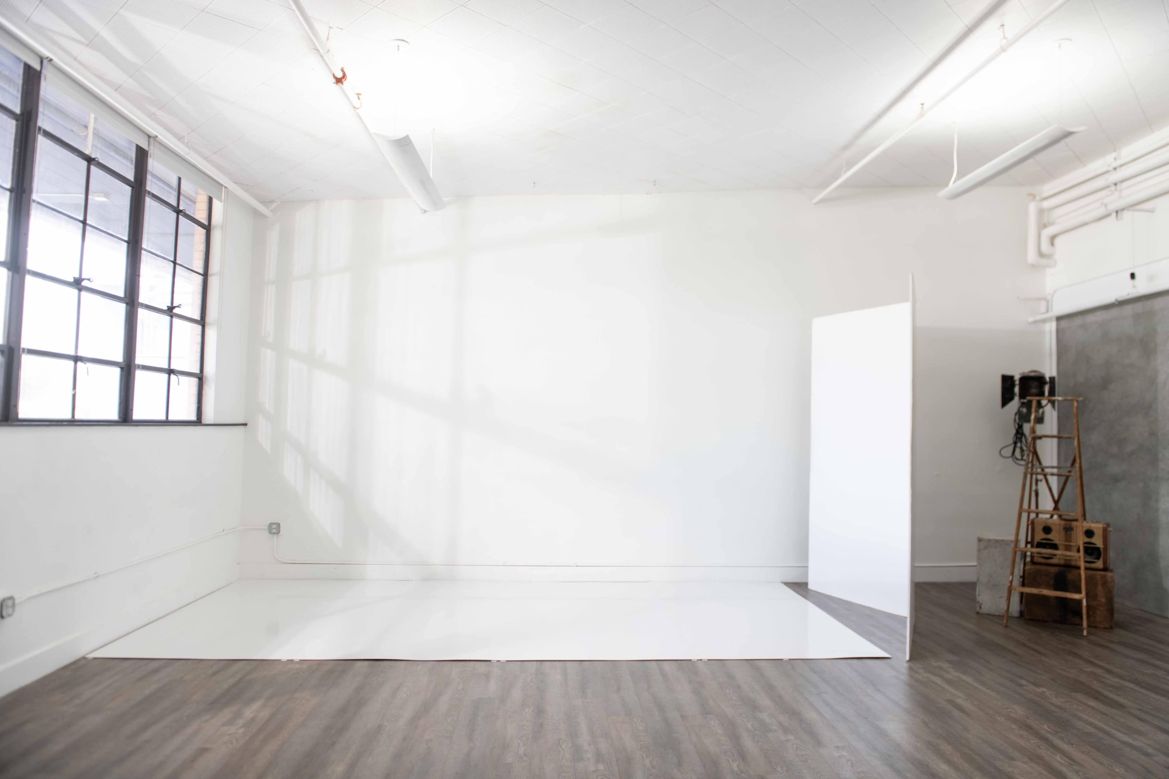 A spacious, empty room with a white wall and a large white floor panel, illuminated by natural light coming through large windows.