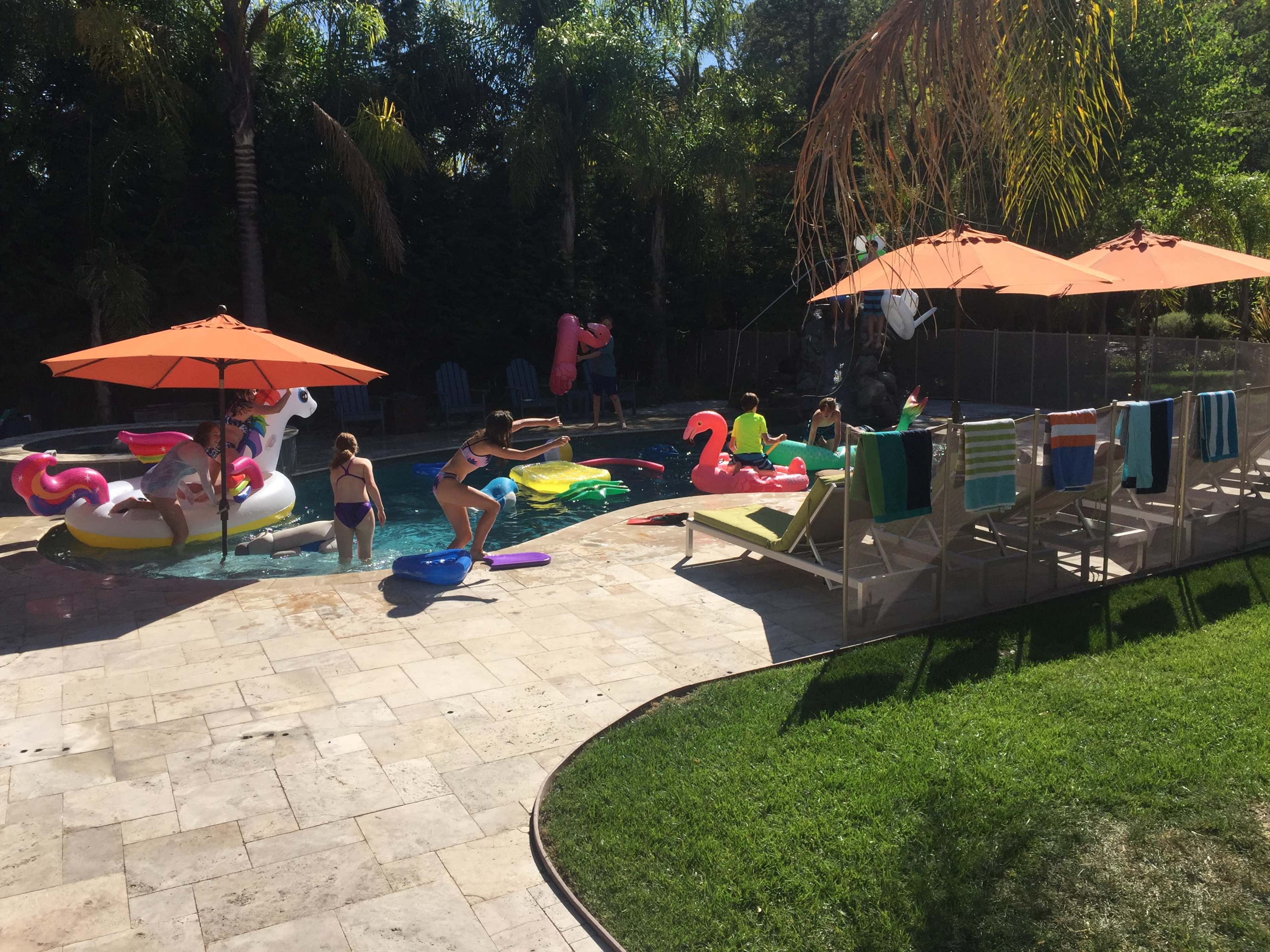 A group of children enjoys playing in a backyard pool surrounded by colorful floaties, lounge chairs, and sun umbrellas.