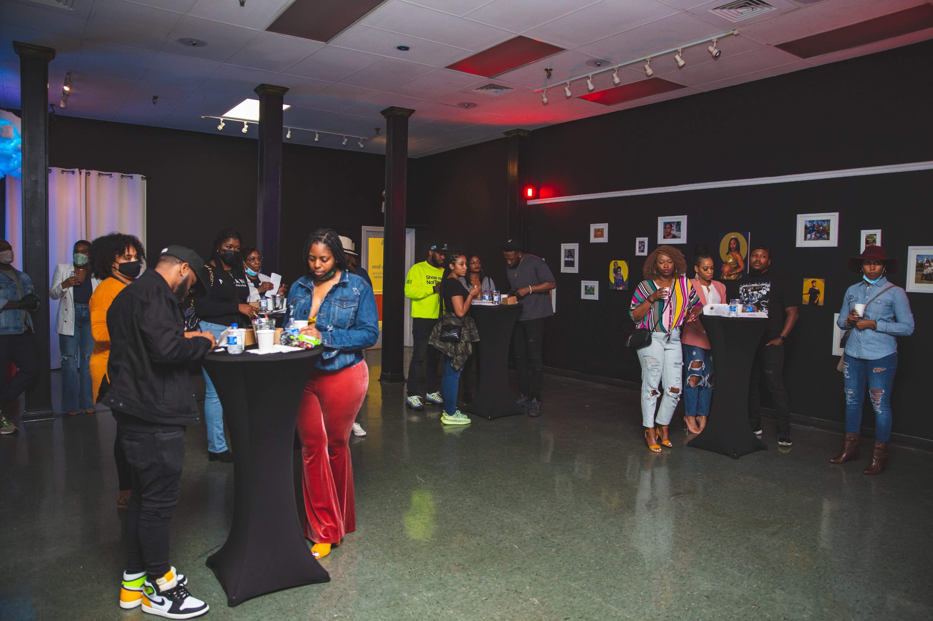 A group of people socializes at tall tables in a dimly lit event space with black walls and colorful artwork.