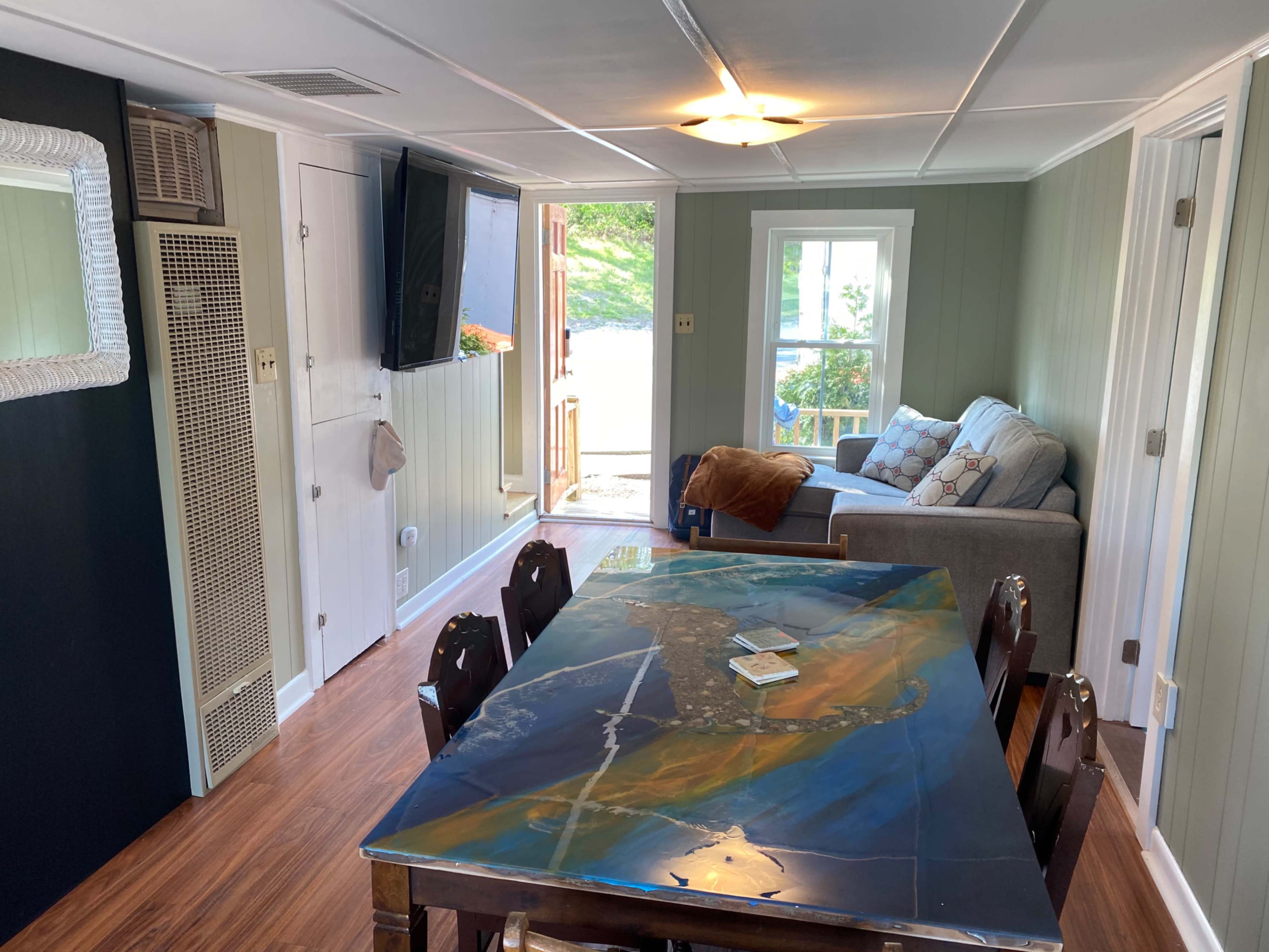 A living space with a dining table in the foreground, a sofa and television in the background, and a doorway leading outside.