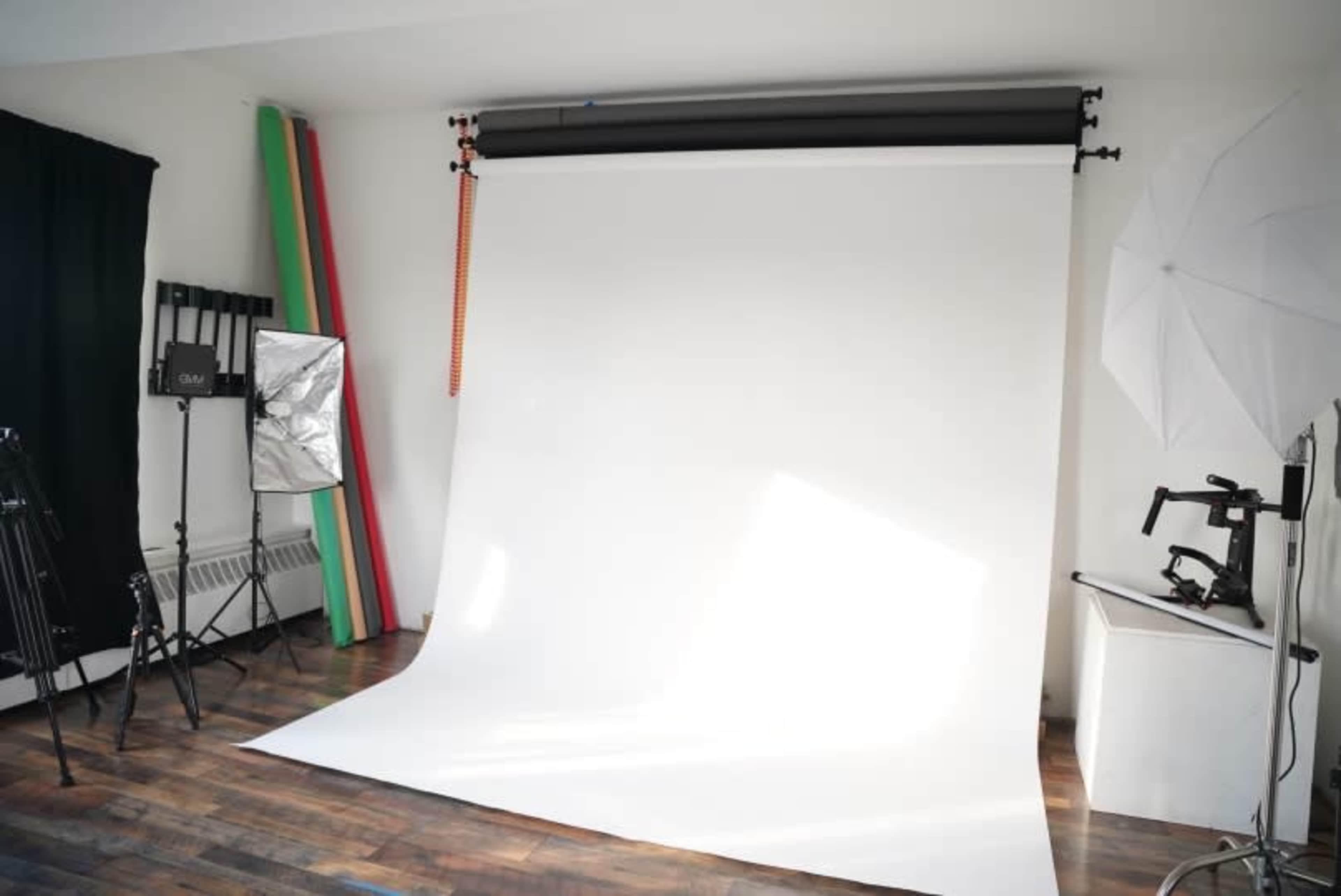 A photography studio with a white backdrop, various lighting equipment, and colored backdrop rolls against the wall.
