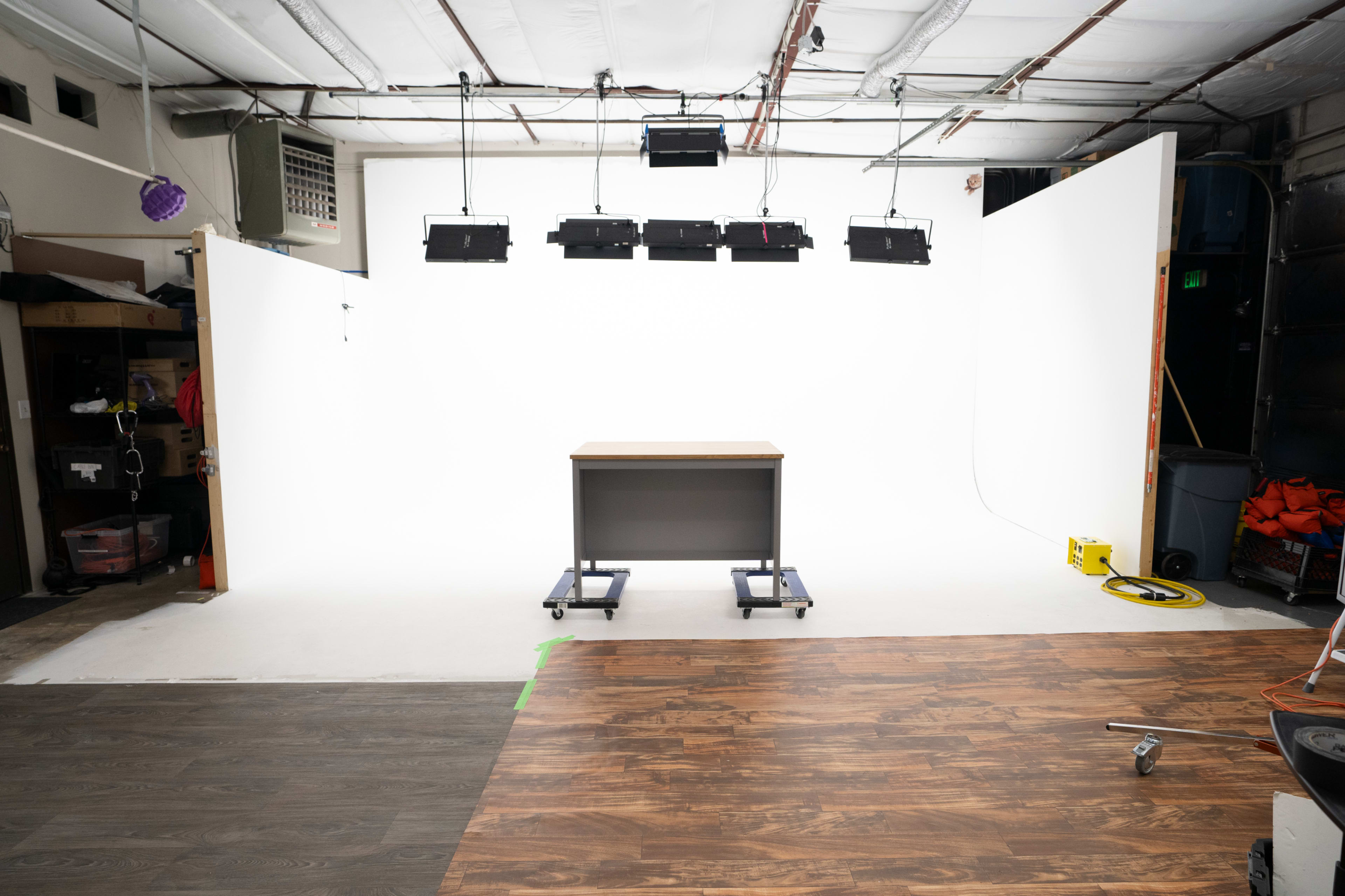 The image shows a minimalistic studio space with a wooden table on wheels, surrounded by a large white backdrop and overhead lighting.