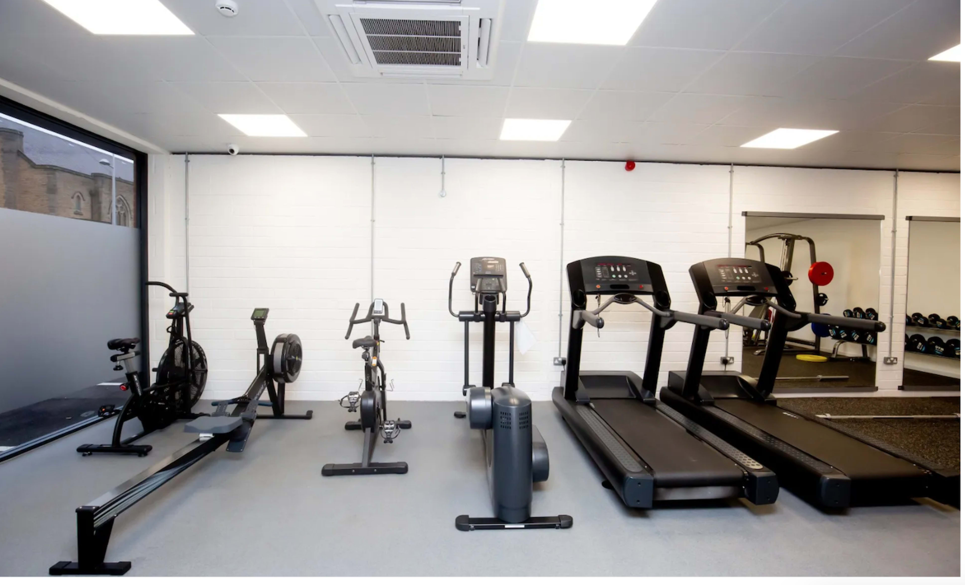 The image shows a fitness area with various exercise machines, including treadmills, an elliptical, a stationary bike, and a rowing machine, along with a set of free weights in the background.