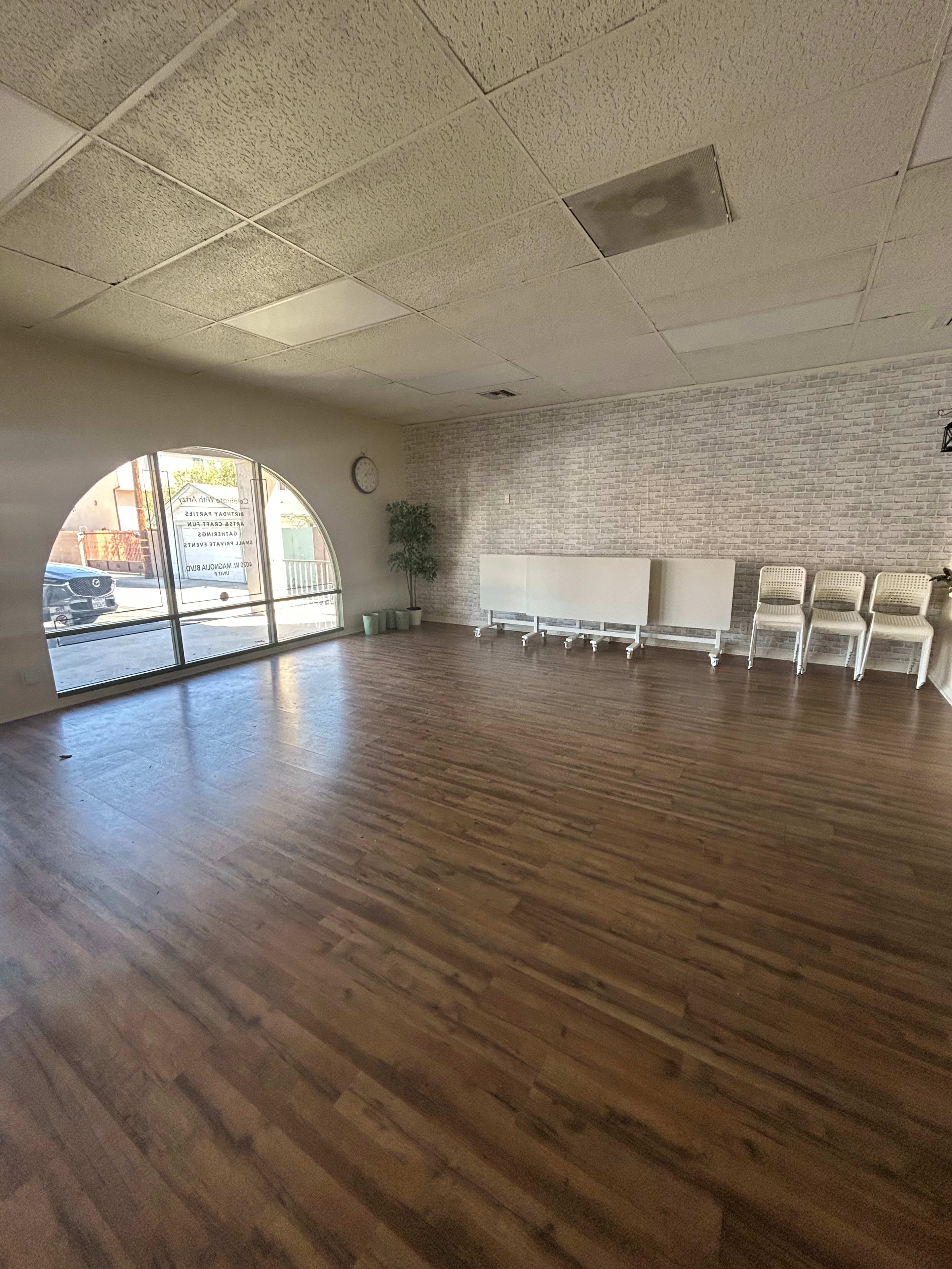 The image shows a spacious, empty room with wooden flooring, a large arched window, and several white chairs lined against the wall.