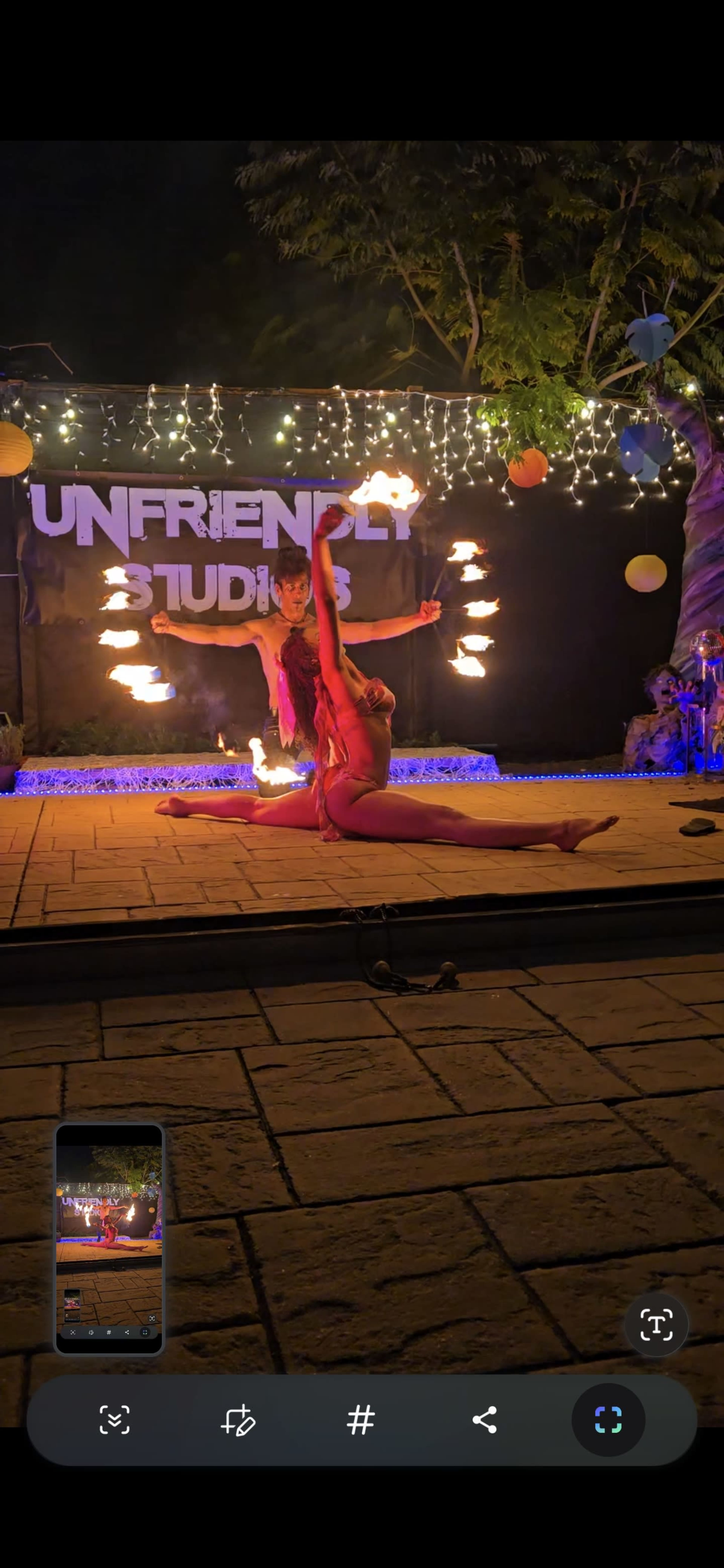 A performer in a pink outfit showcases a split pose while holding a flaming object, with another performer in the background also handling fire, illuminated by string lights and colorful decorations.