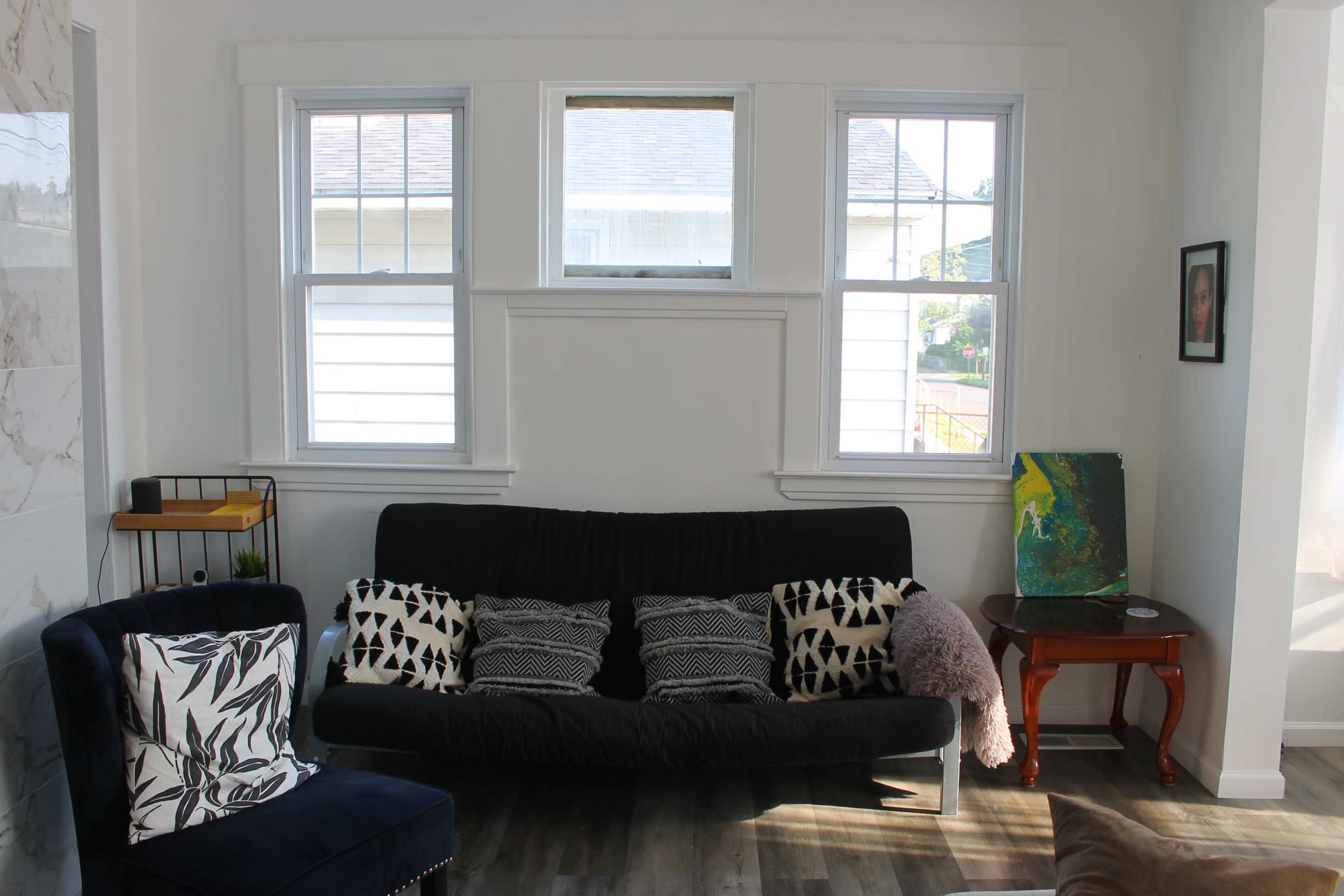 A living room with a black futon, patterned cushions, and two windows providing natural light.