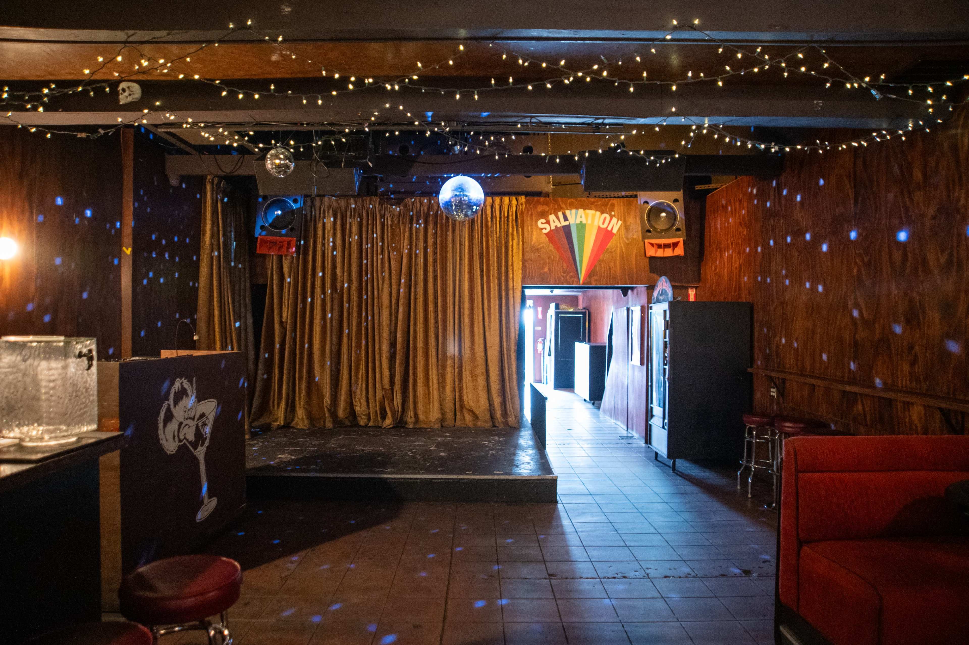The interior of a dimly lit nightclub features a disco ball, a small stage with gold curtains, and red seating along the sides.