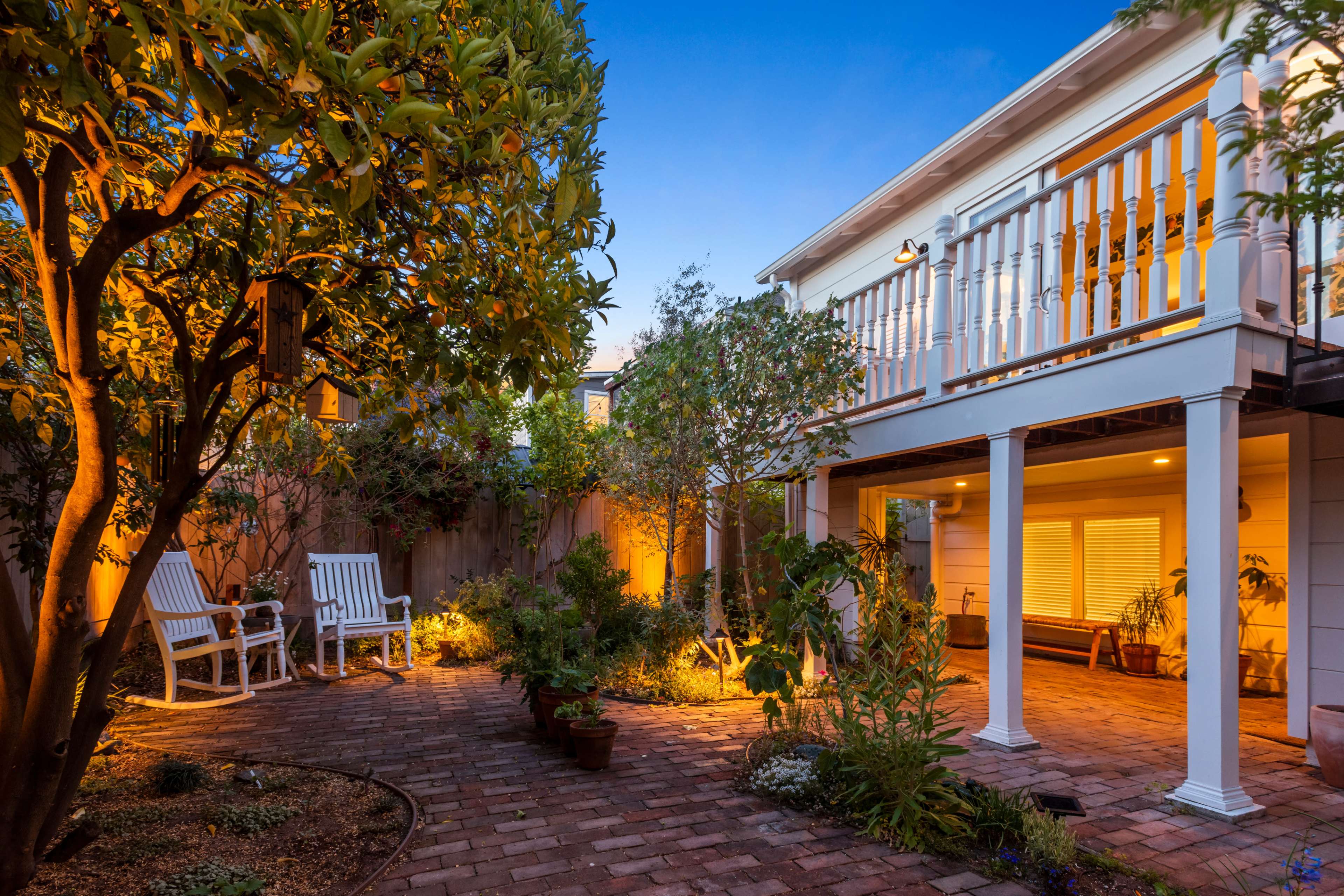 A brick-paved backyard with garden plants, two white chairs, and a lit area beneath a deck.