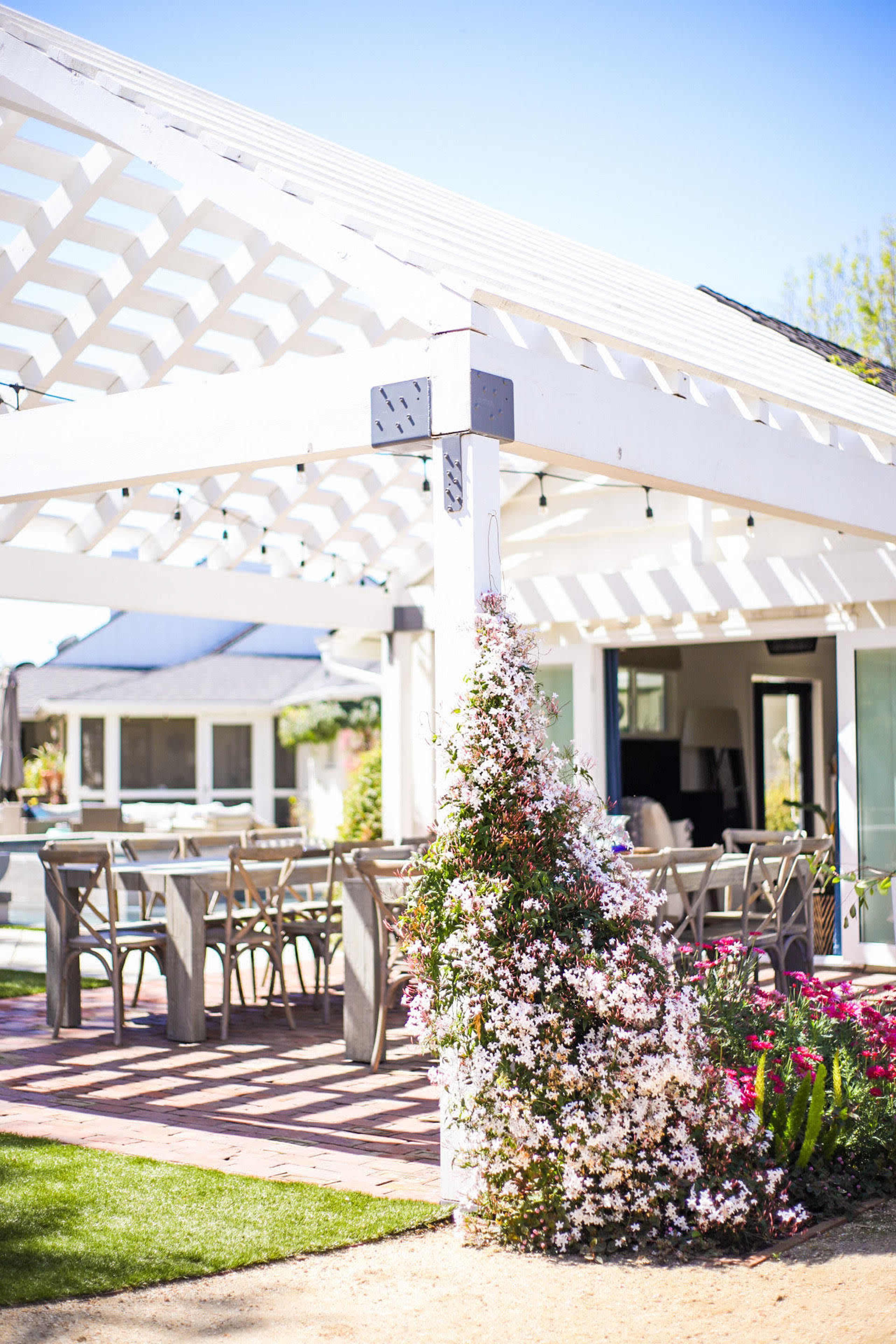 A pergola with a white trellis structure provides shade over a dining area, surrounded by flowering plants in a landscaped garden.