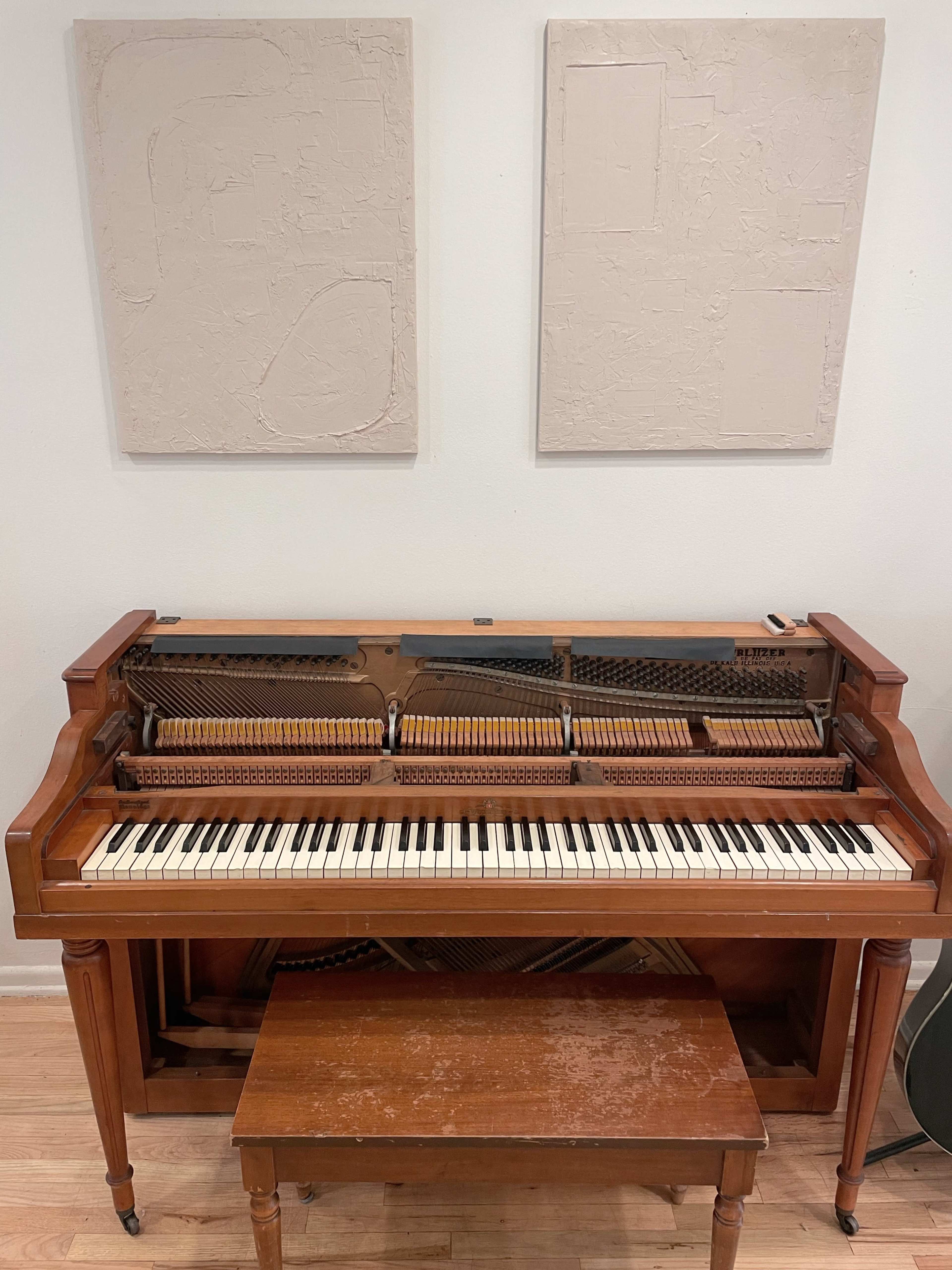 A wooden piano with an open top and a small bench sits in front of two textured beige wall panels.