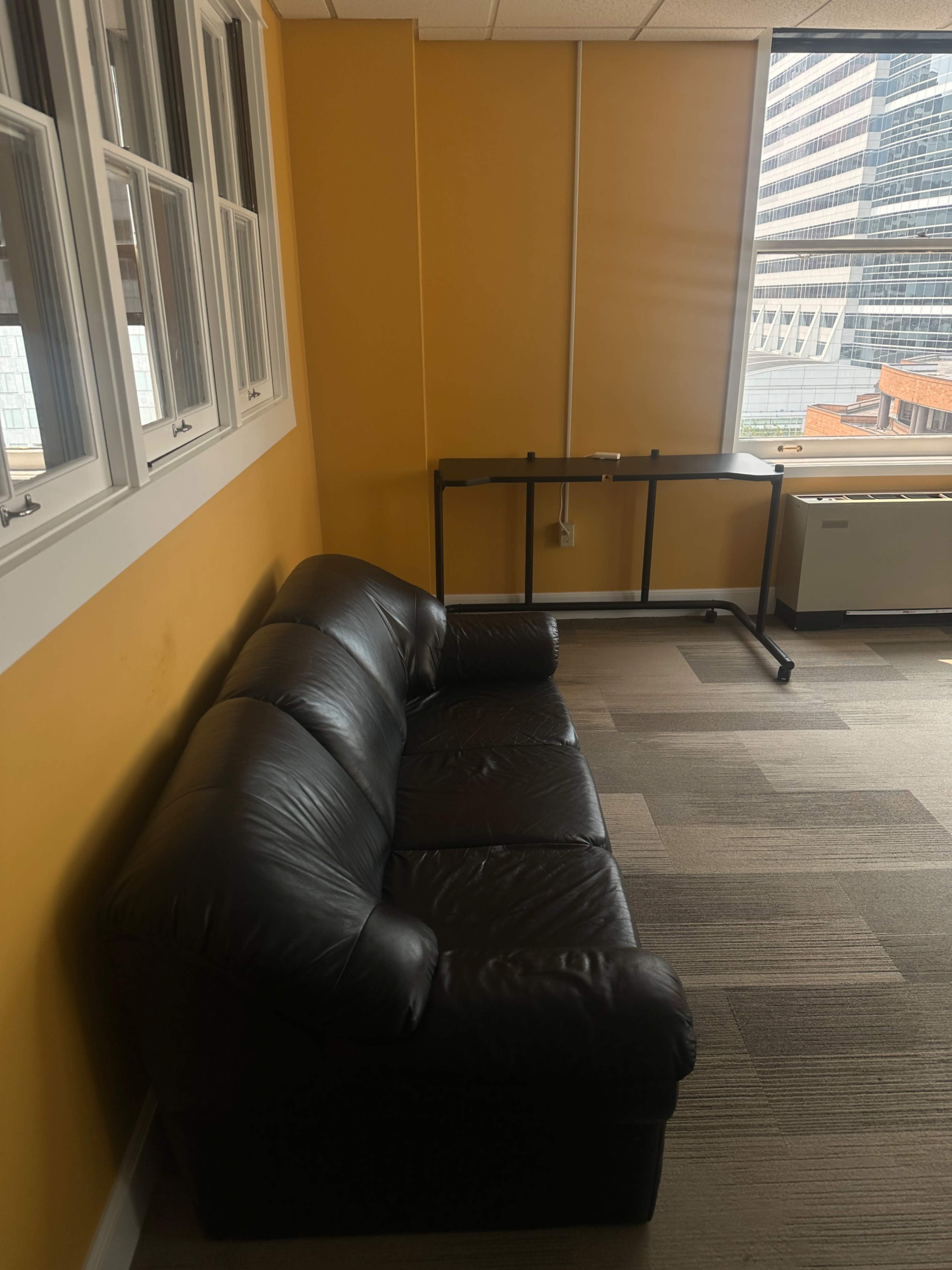 The image shows a black leather couch positioned against a yellow wall in a room with large windows and a desk nearby.