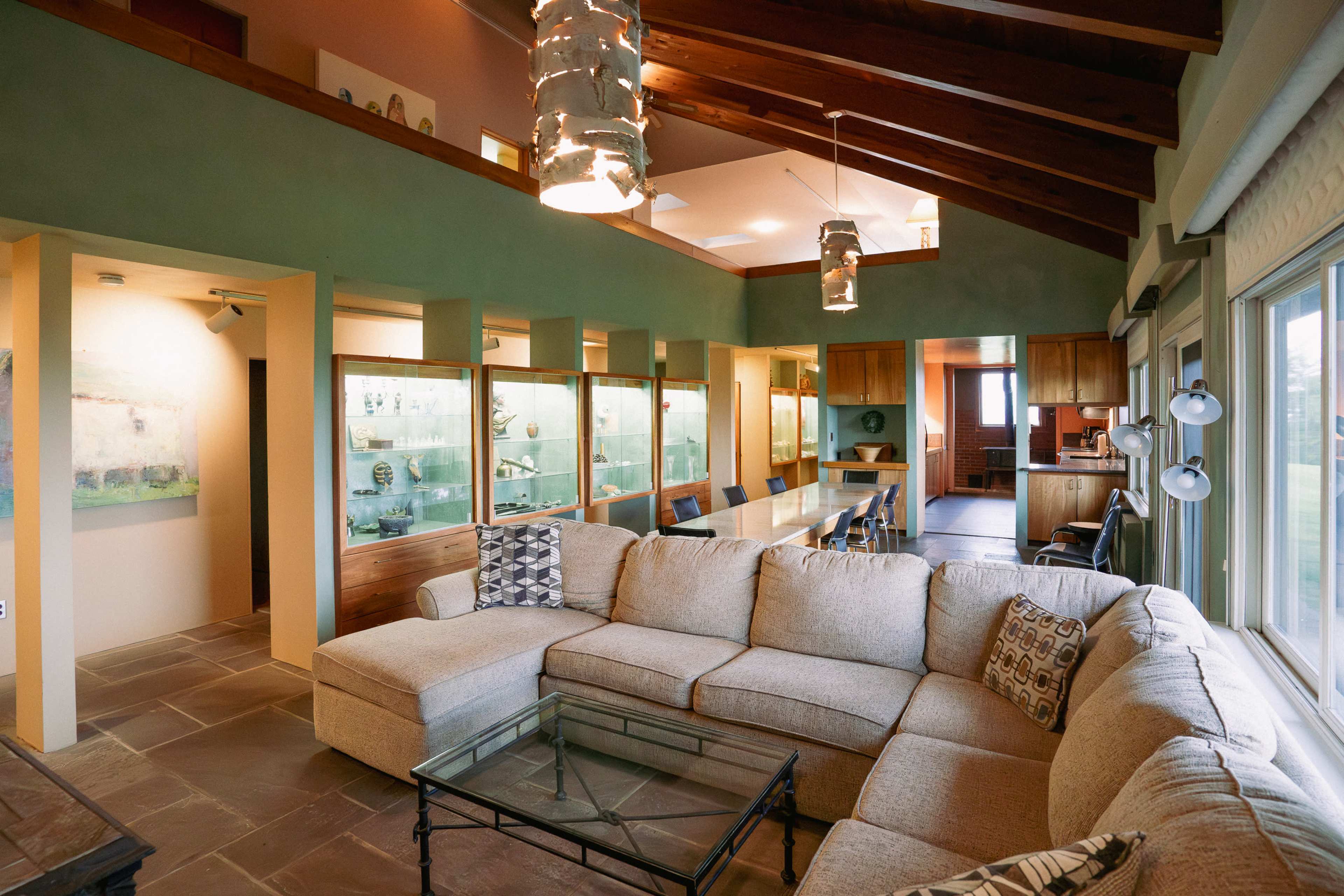 A spacious living area with a large sectional sofa, high wooden ceilings, and an open layout leading to a kitchen and dining area.