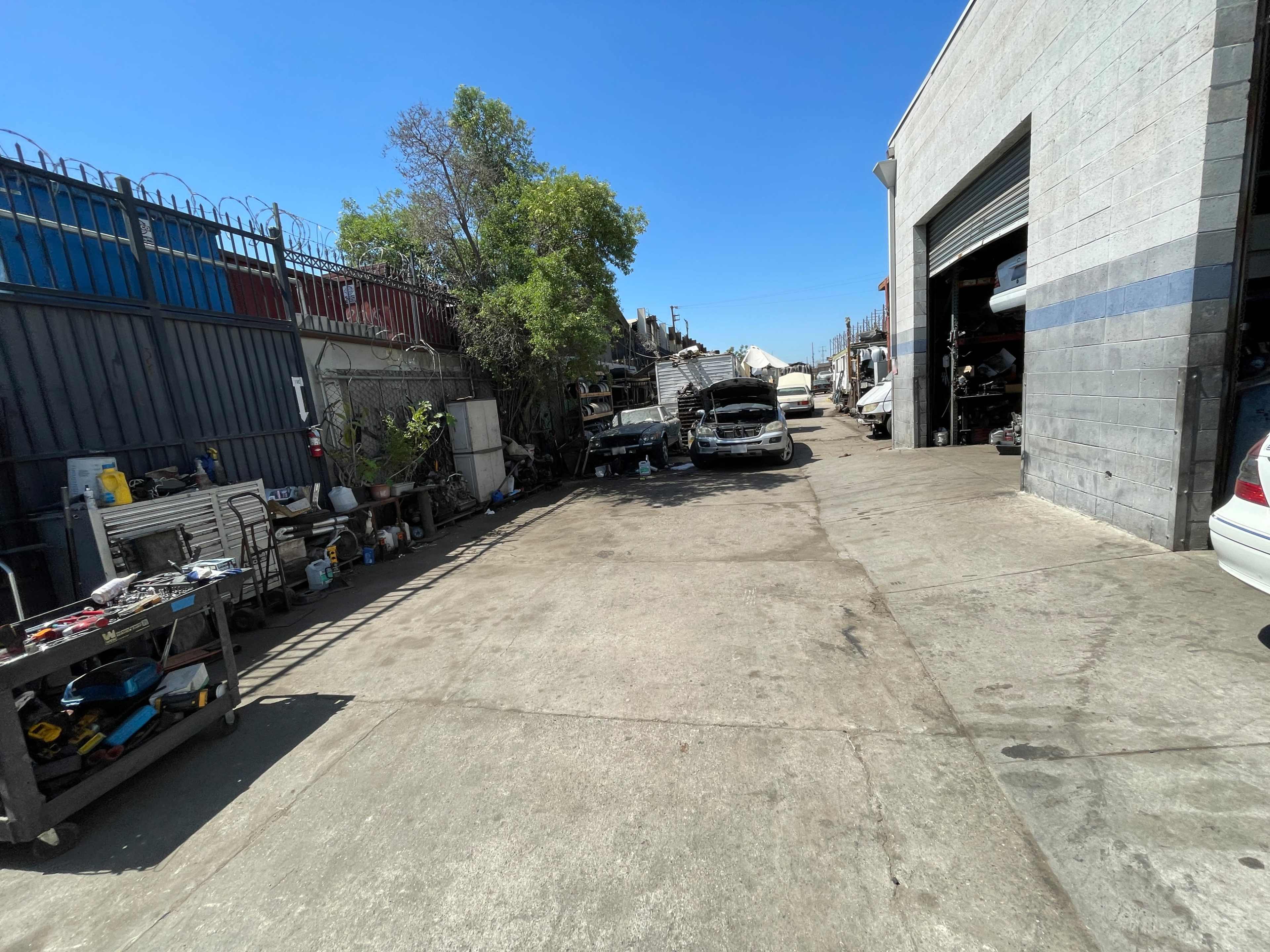 The image shows a vehicle repair yard with several cars parked on the concrete surface, surrounded by tools and equipment.