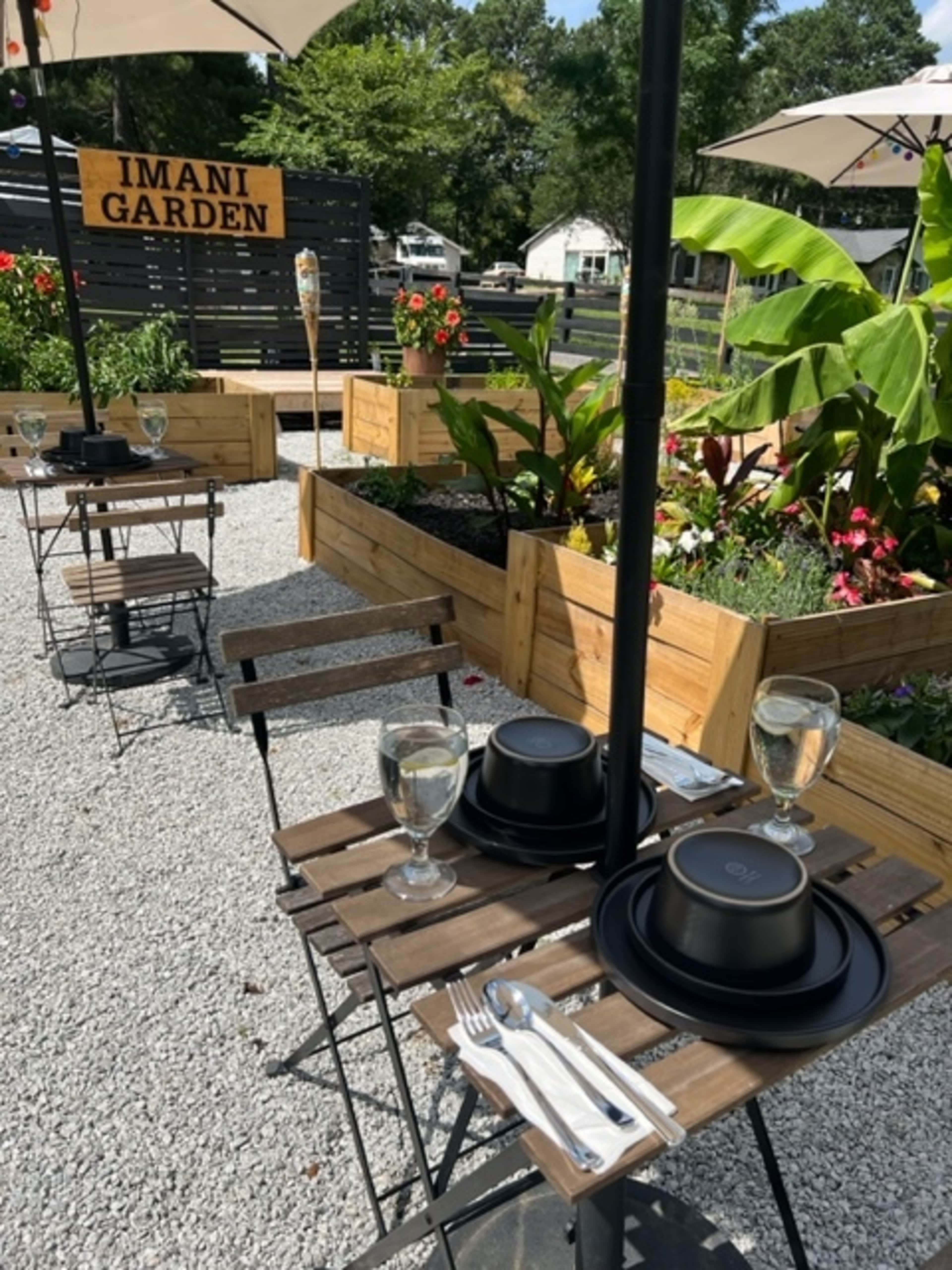 The image shows an outdoor dining area at Imani Garden, featuring tables with black plates and glasses, surrounded by raised garden beds filled with flowers.