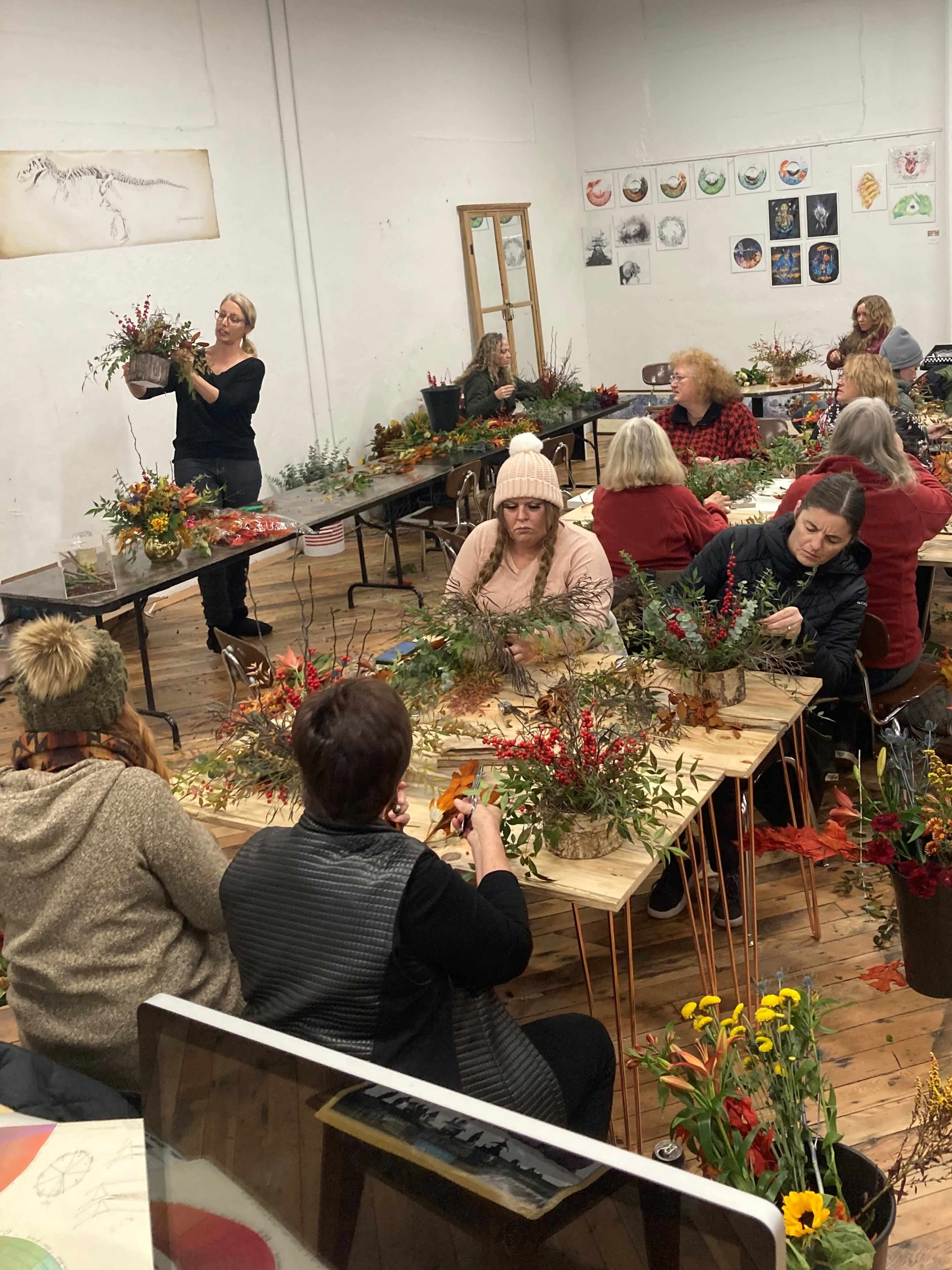 A group of people gathers around tables in a spacious room, engaged in floral arranging with a variety of plants and decorations.