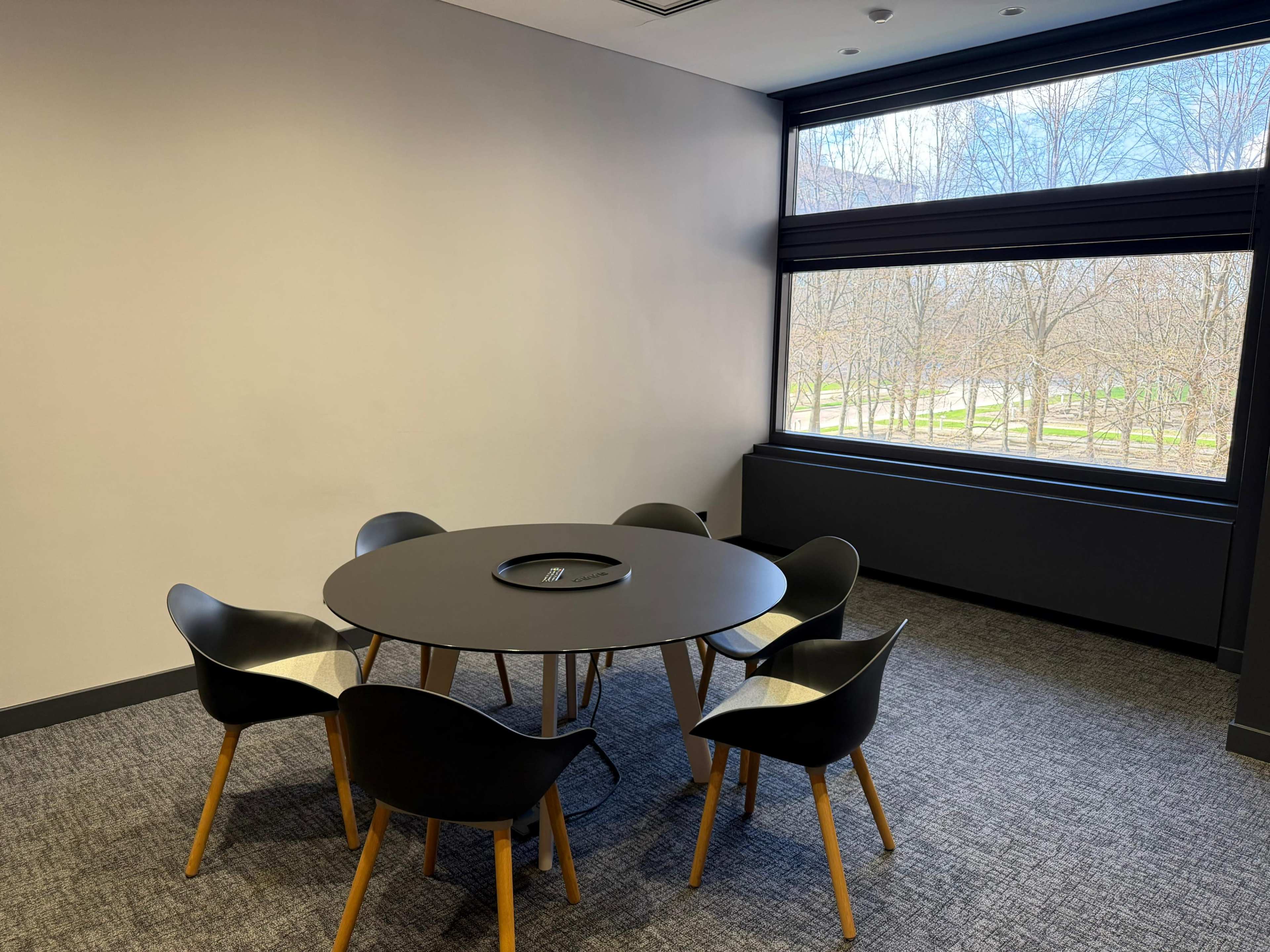 A round table with six chairs is set up in a modern conference room that features a large window overlooking a landscape with bare trees.