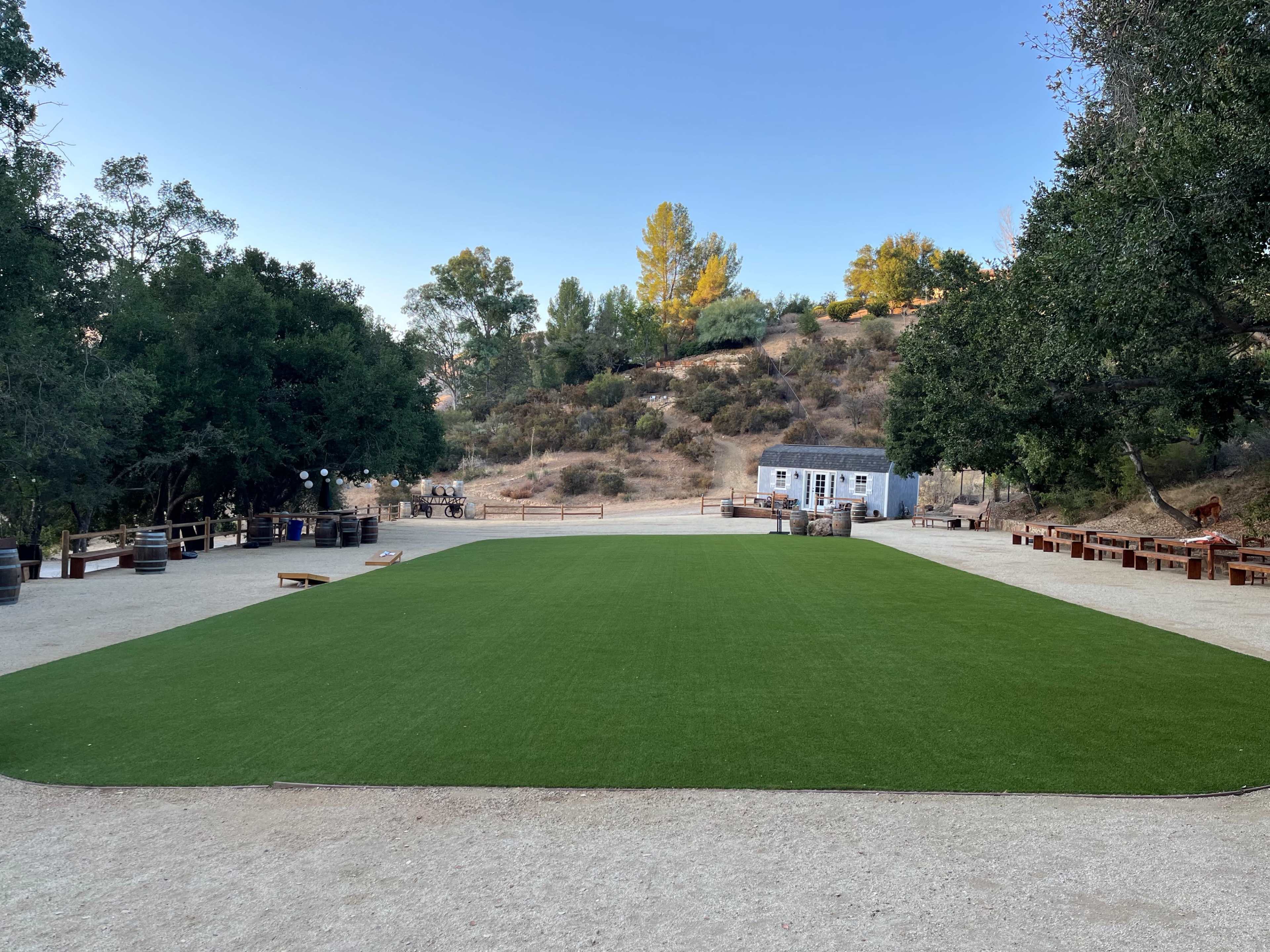 A rectangular area of artificial grass is surrounded by trees and rustic wooden furniture in a rural setting.
