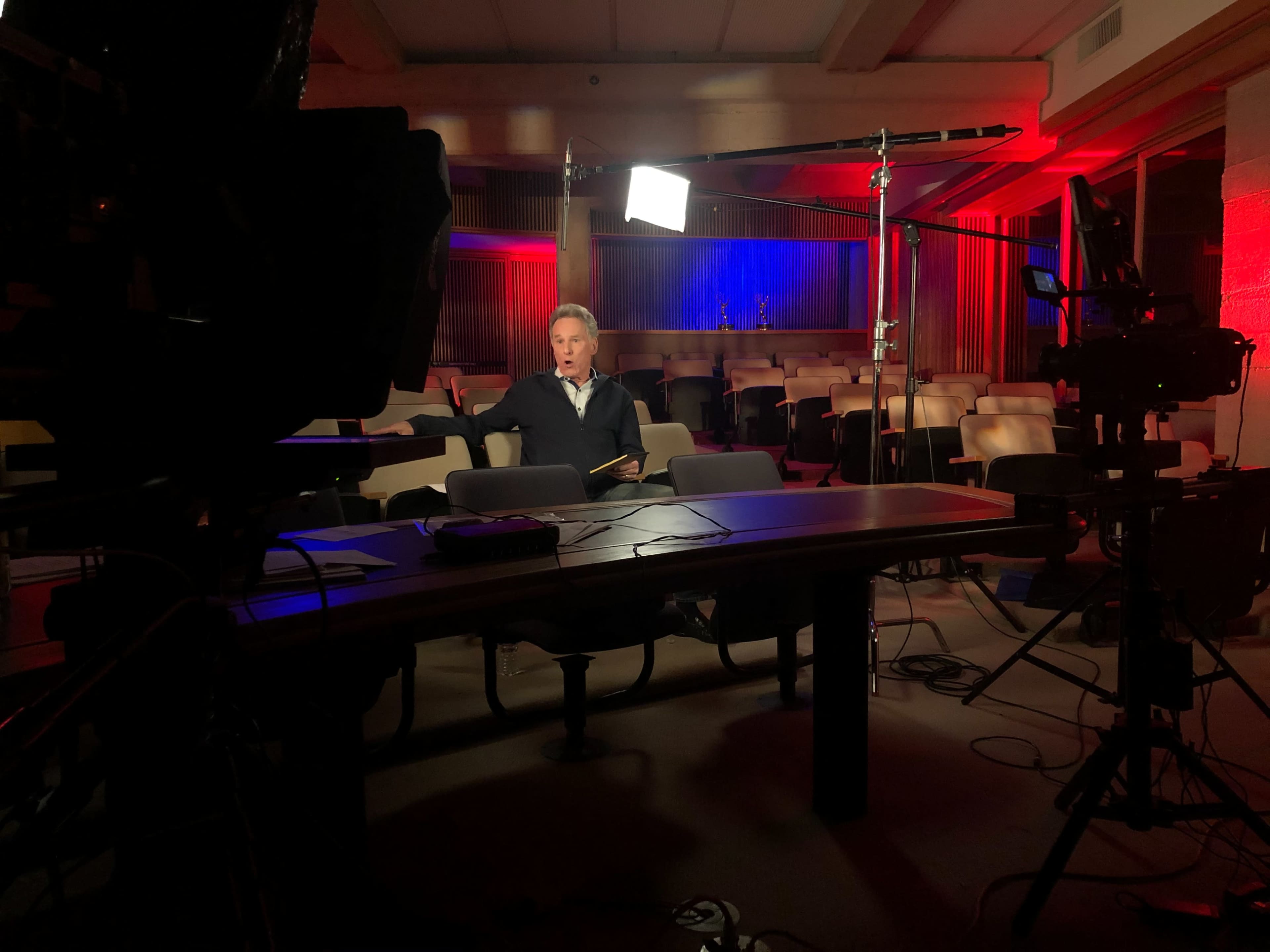 A man in formal attire sits at a table in a dimly lit room with rows of empty chairs, while camera equipment is set up around him.