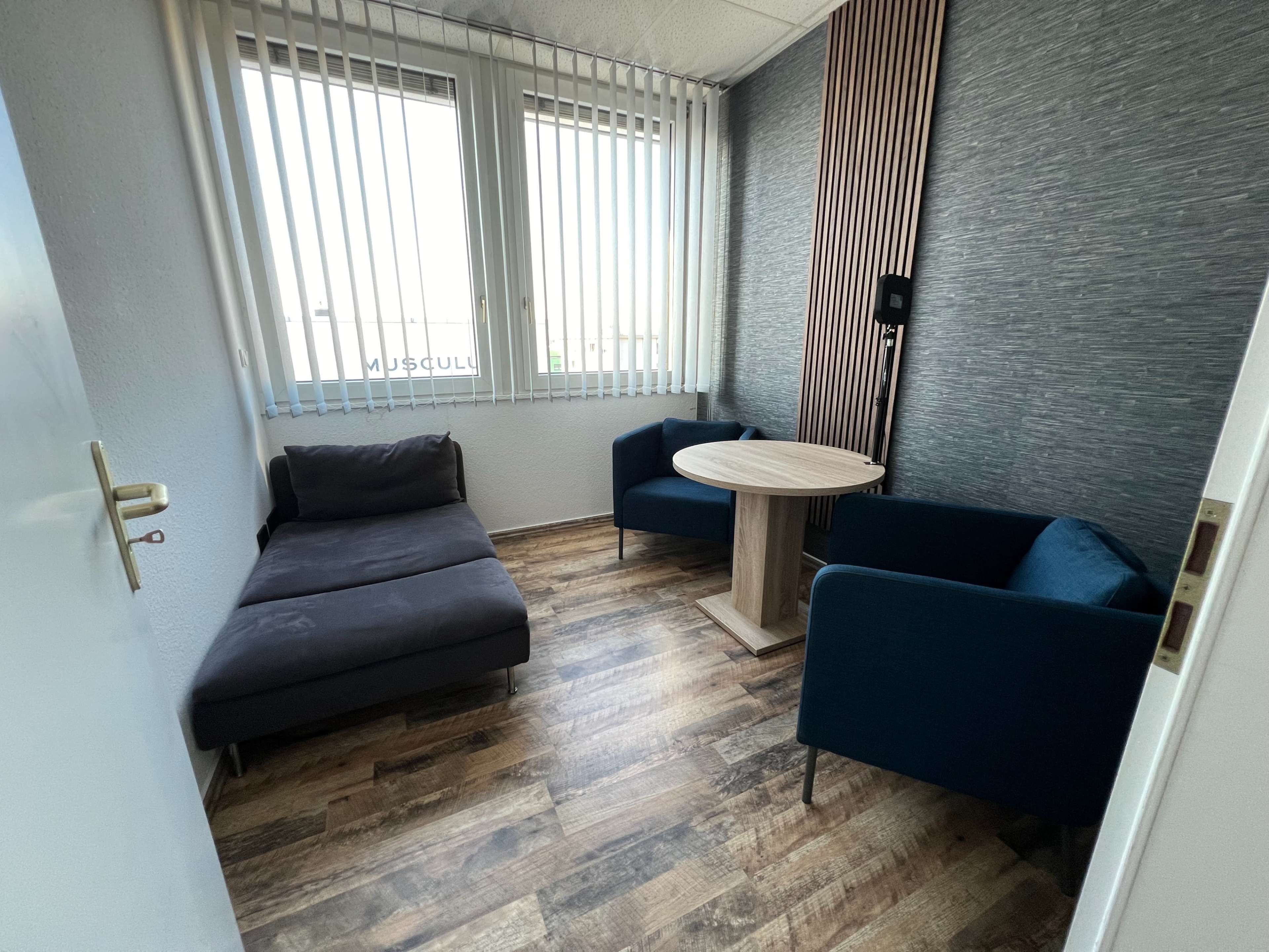 The image shows a small waiting room with a dark sofa, two blue chairs, and a round wooden table, featuring large windows with vertical blinds.