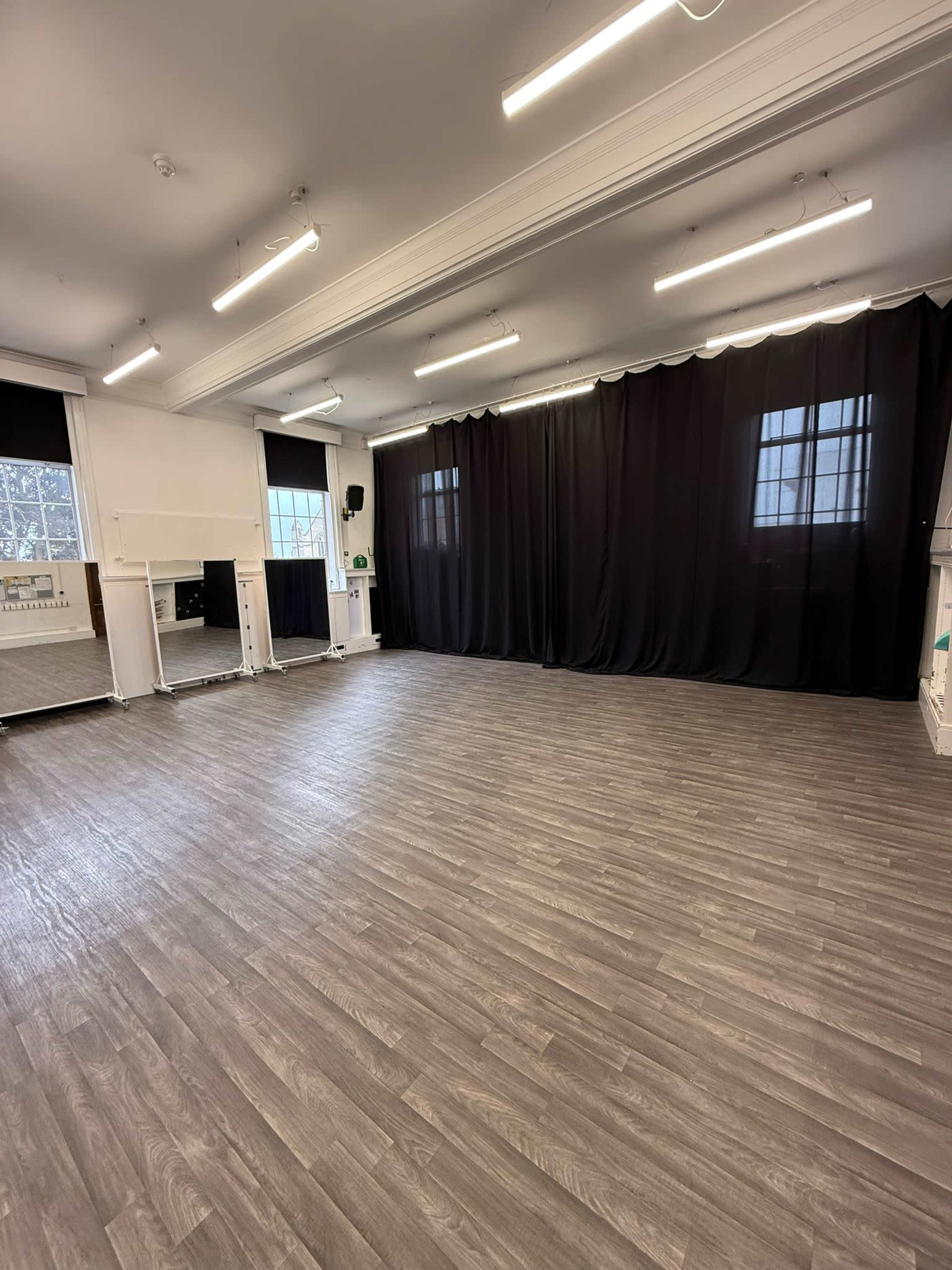 The image shows an empty studio space with wooden flooring, black curtains, and large mirrors on one wall.