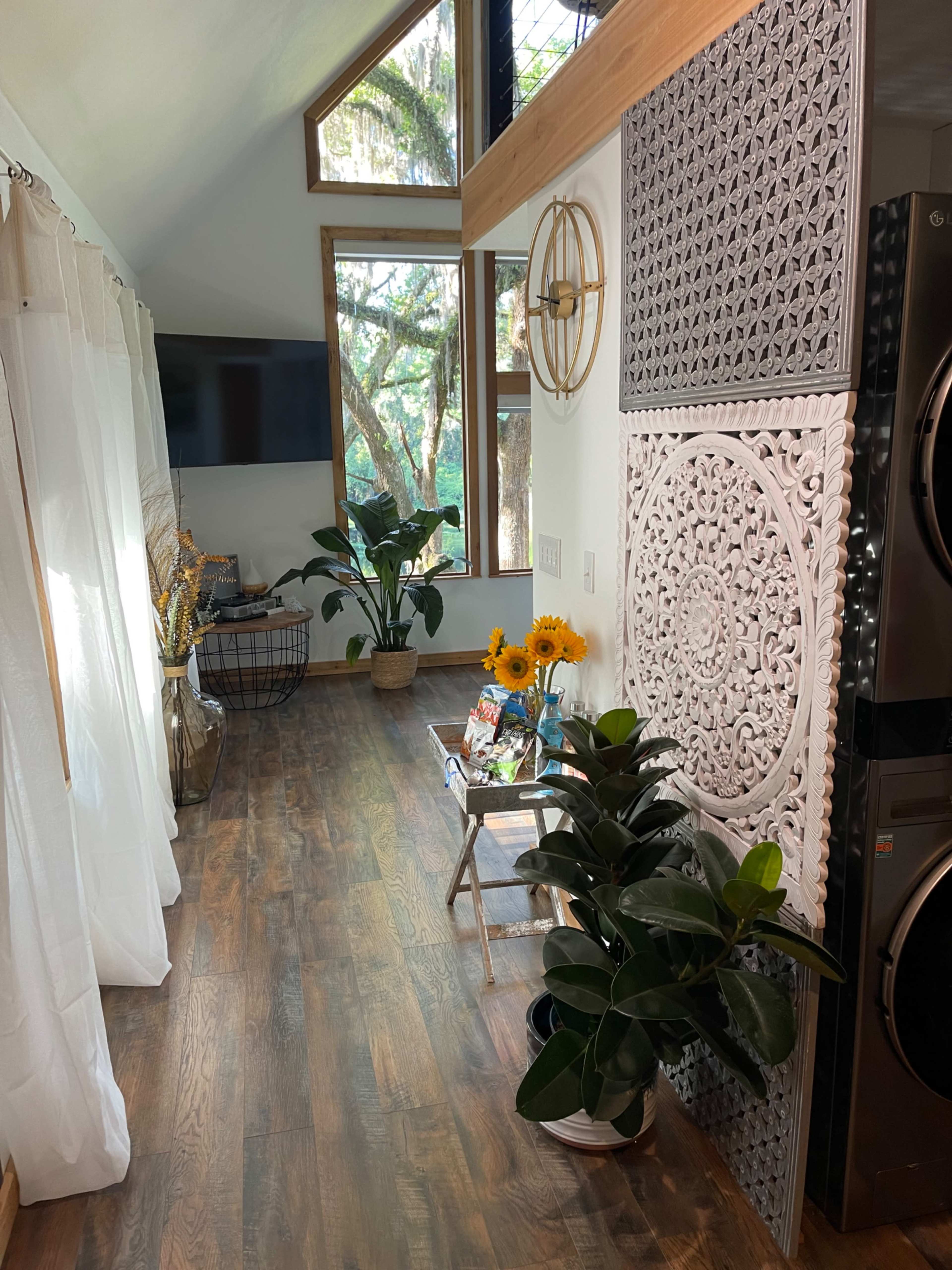 The image shows a bright interior space with plants, a large wall decoration, a television, and a wooden floor.