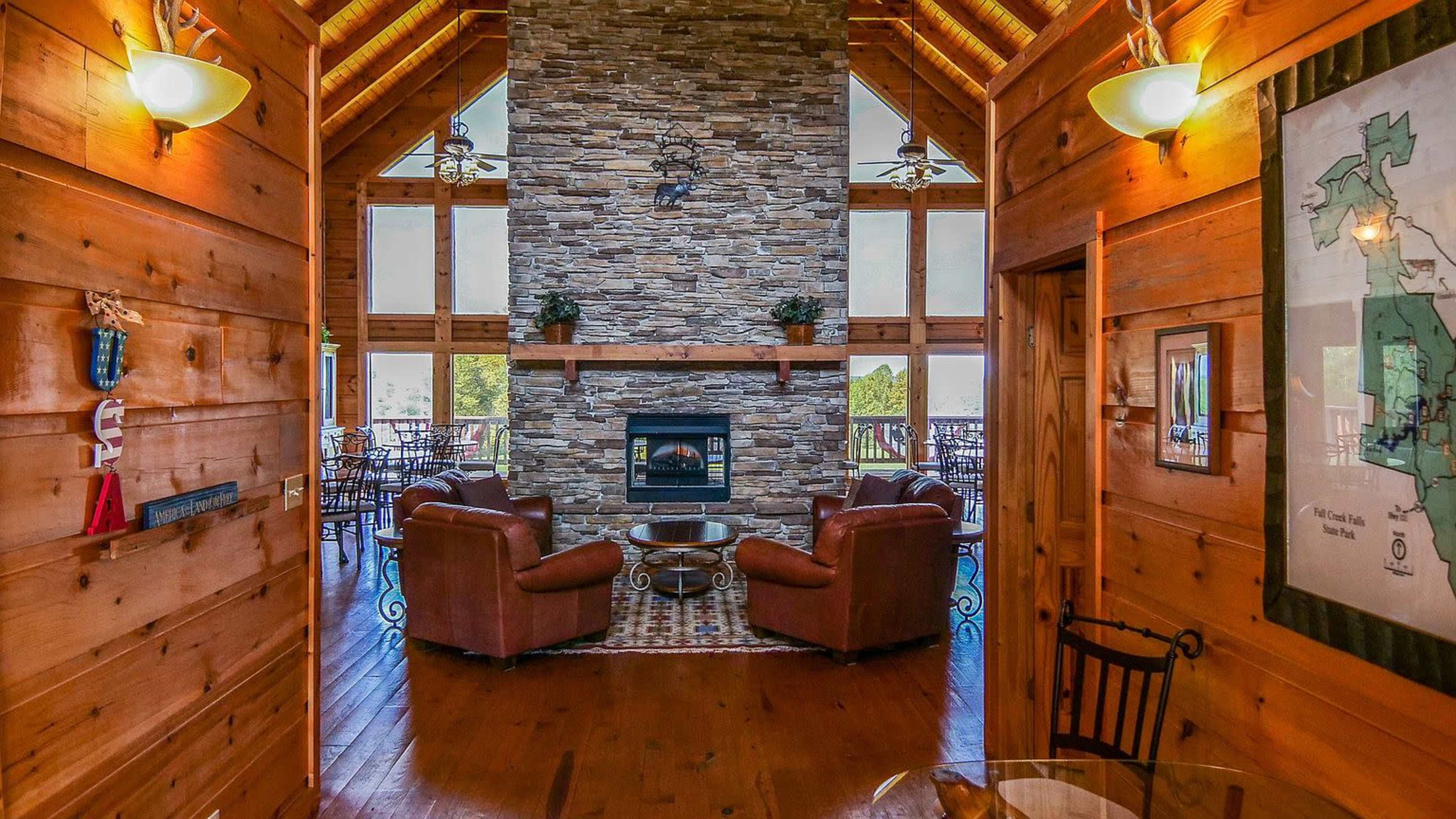 The image shows a wooden interior of a cabin with a stone fireplace at its center, flanked by large windows and wooden furniture.