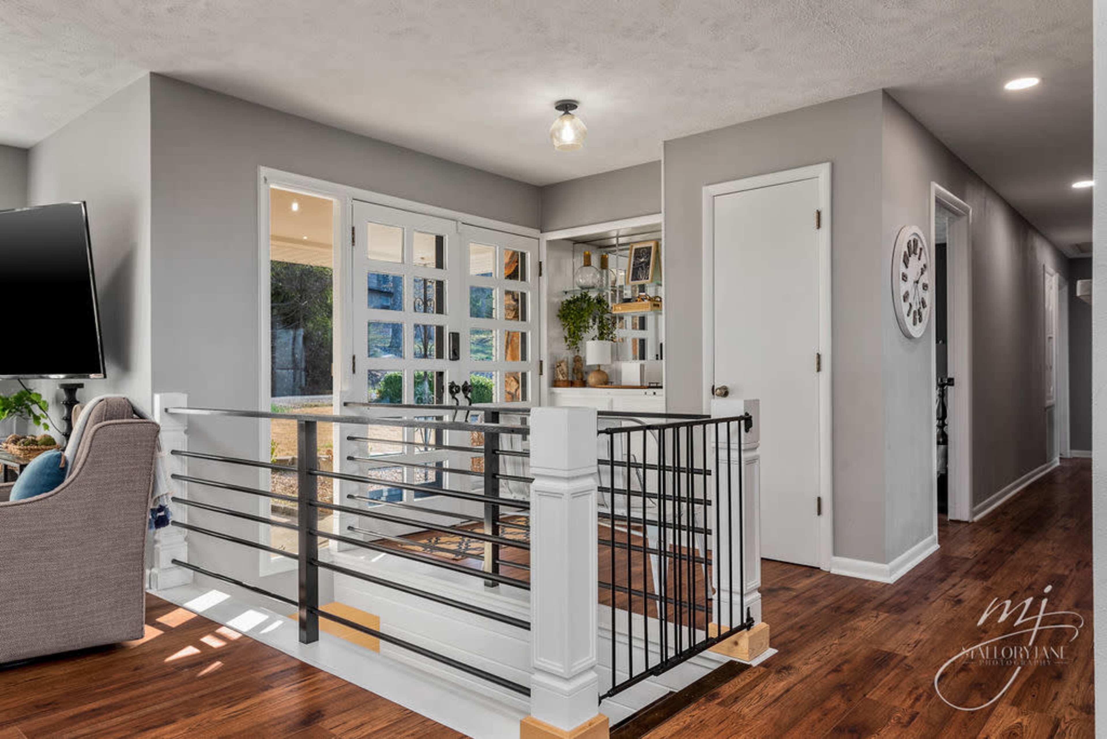 The image shows a modern hallway with a railing, leading to a glass door that opens to a bright room, and features wood flooring and neutral gray walls.