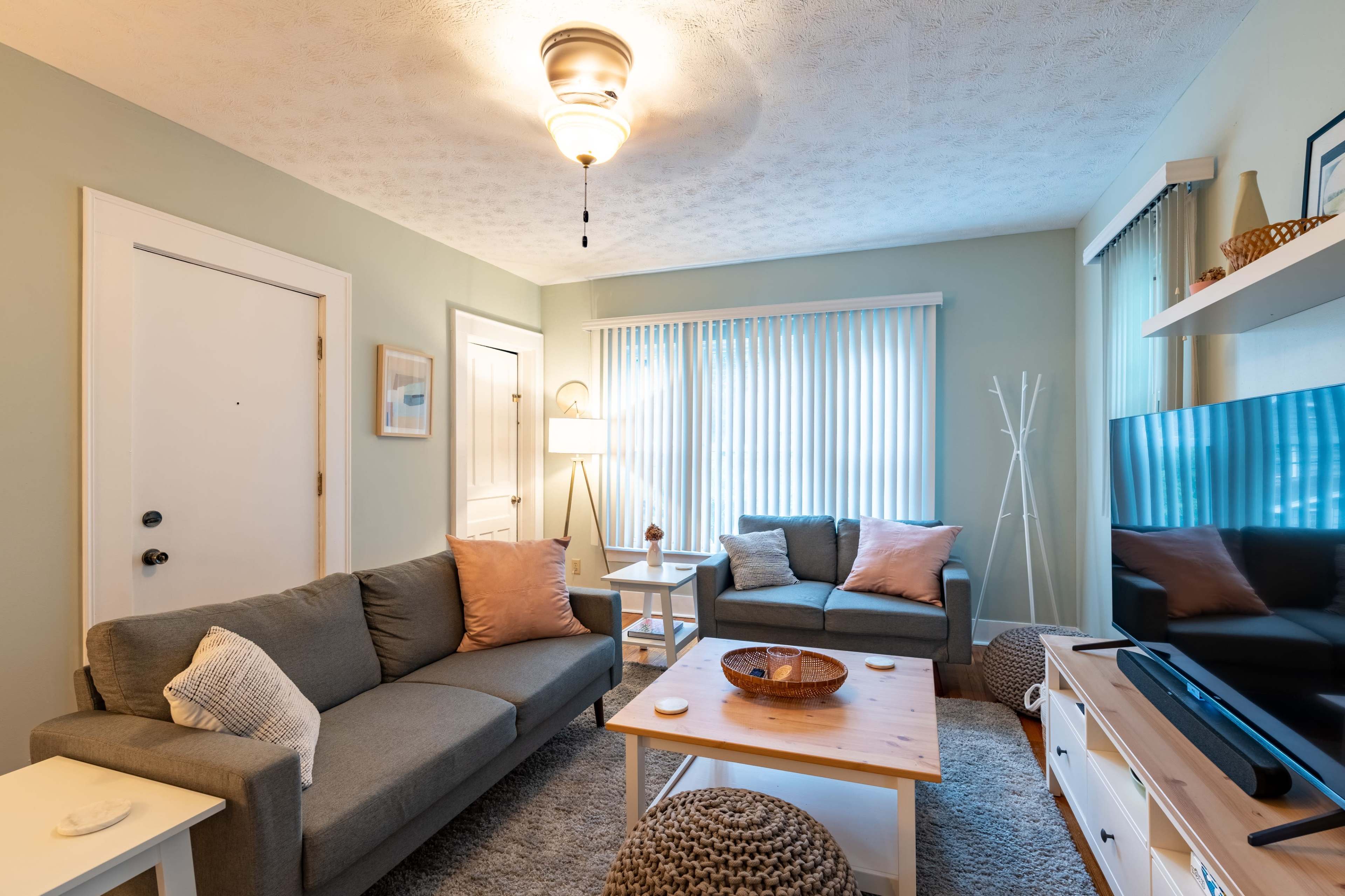 The living room features two gray sofas with pink pillows, a wooden coffee table, and a TV against a light green wall, illuminated by natural light through sheer curtains.