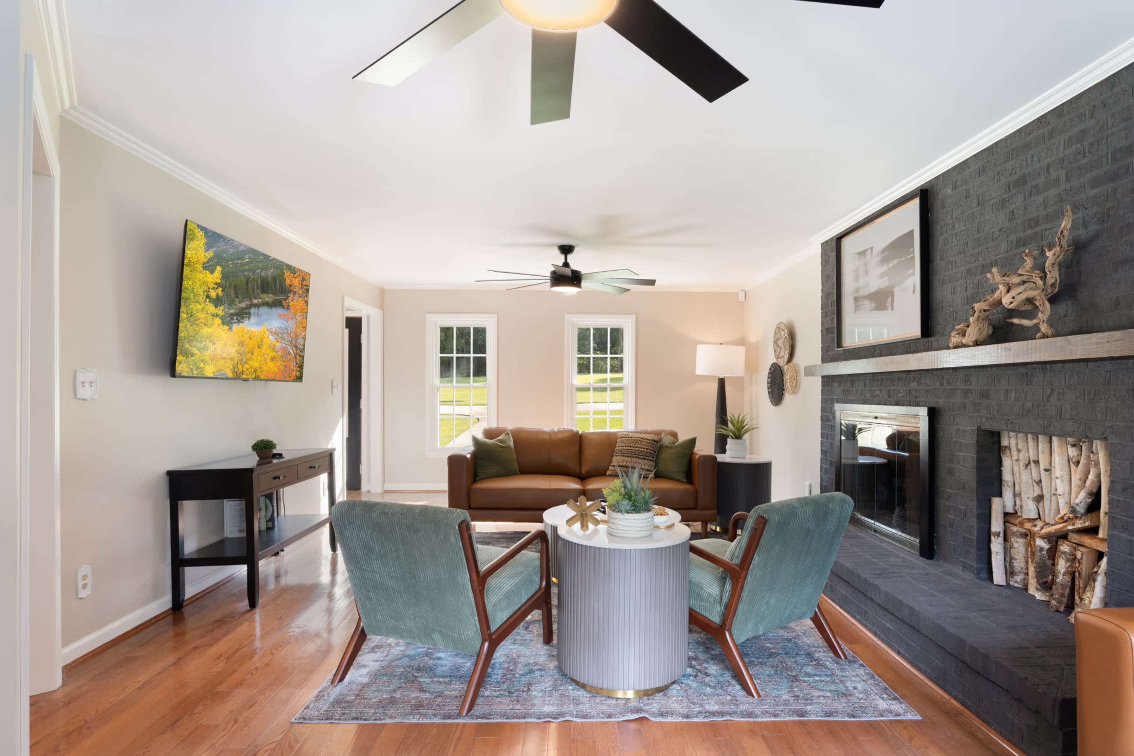 The image shows a living room with a brown sectional sofa, two green velvet chairs, a round coffee table, and a black brick fireplace, all illuminated by natural light from the large windows.
