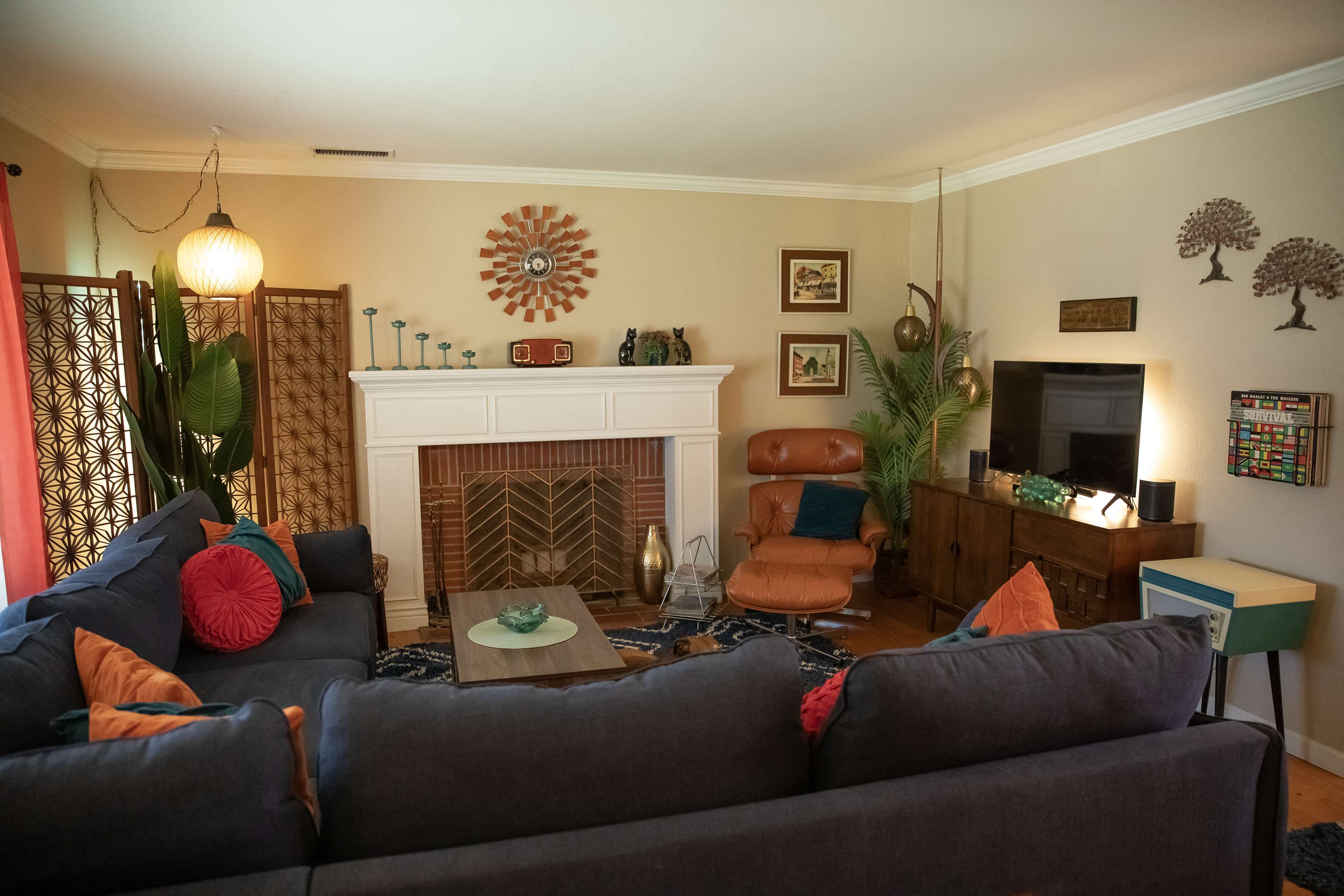 A cozy living room features a sectional sofa, a fireplace with decorative accents, a wooden media console, and various greenery throughout the space.