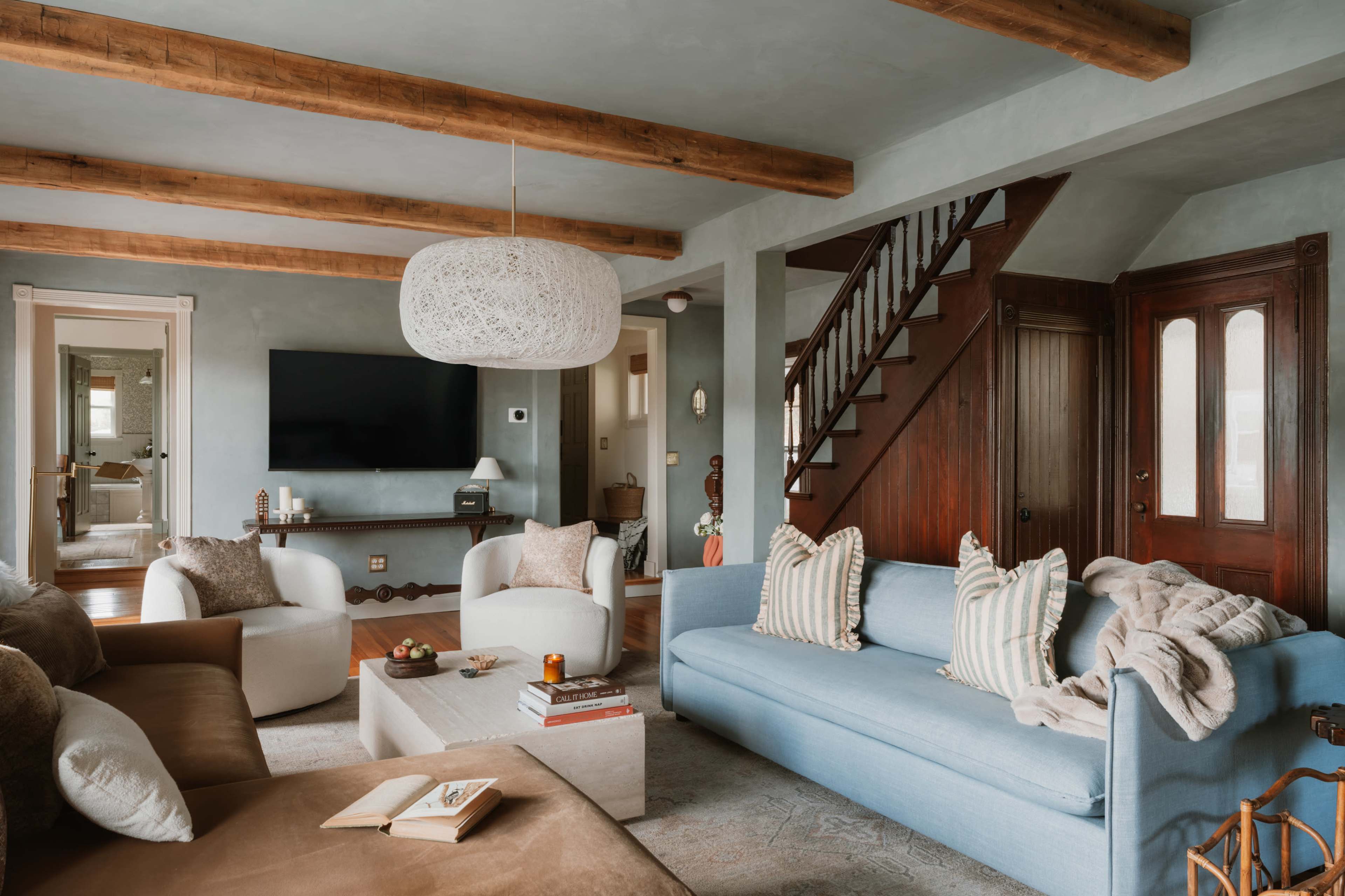 A spacious living room features a mix of light and dark furniture, wooden beams, and a large television mounted on the wall.