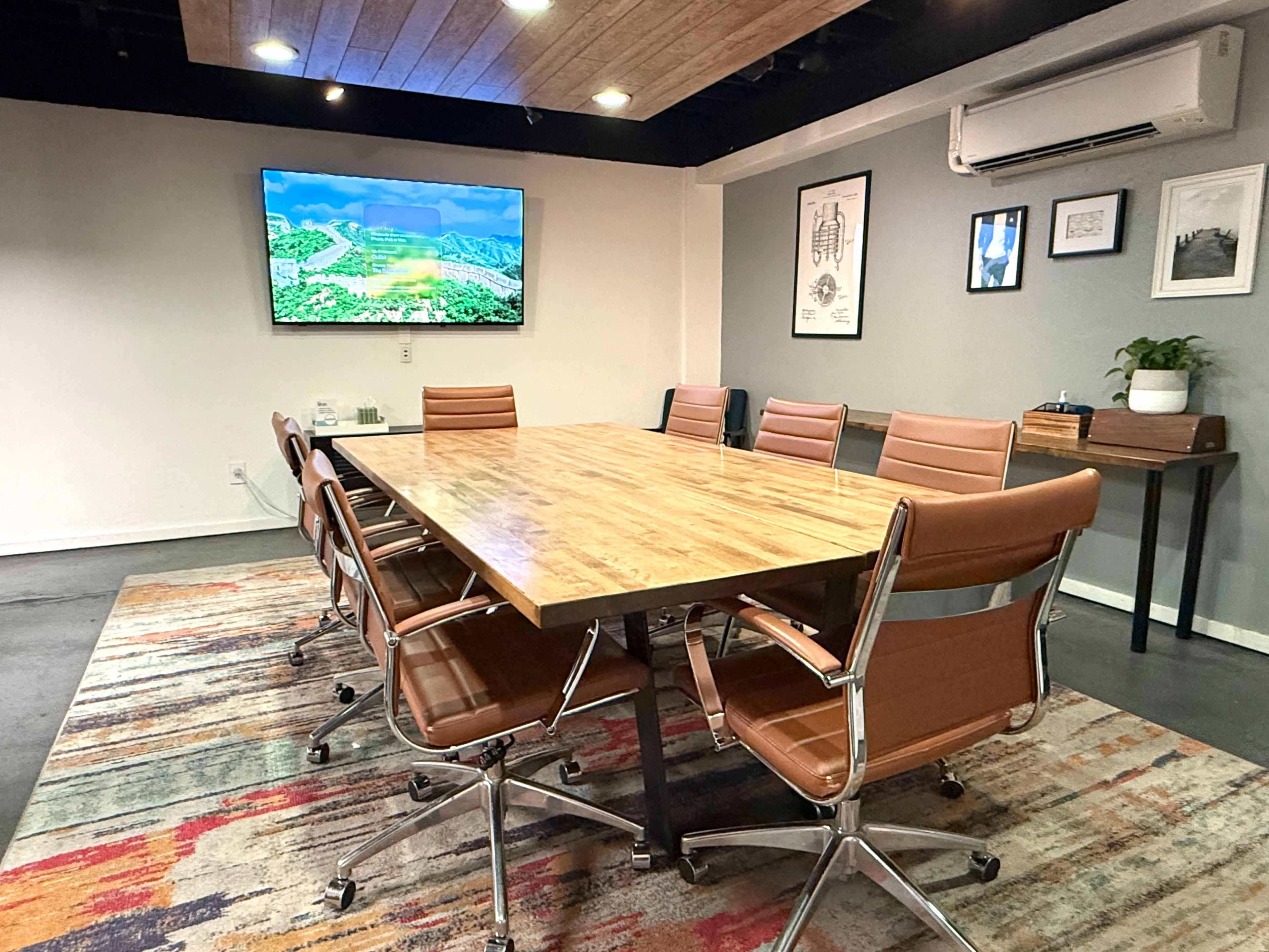 The image shows a modern conference room with a long wooden table, leather chairs, and a wall-mounted screen displaying a landscape.