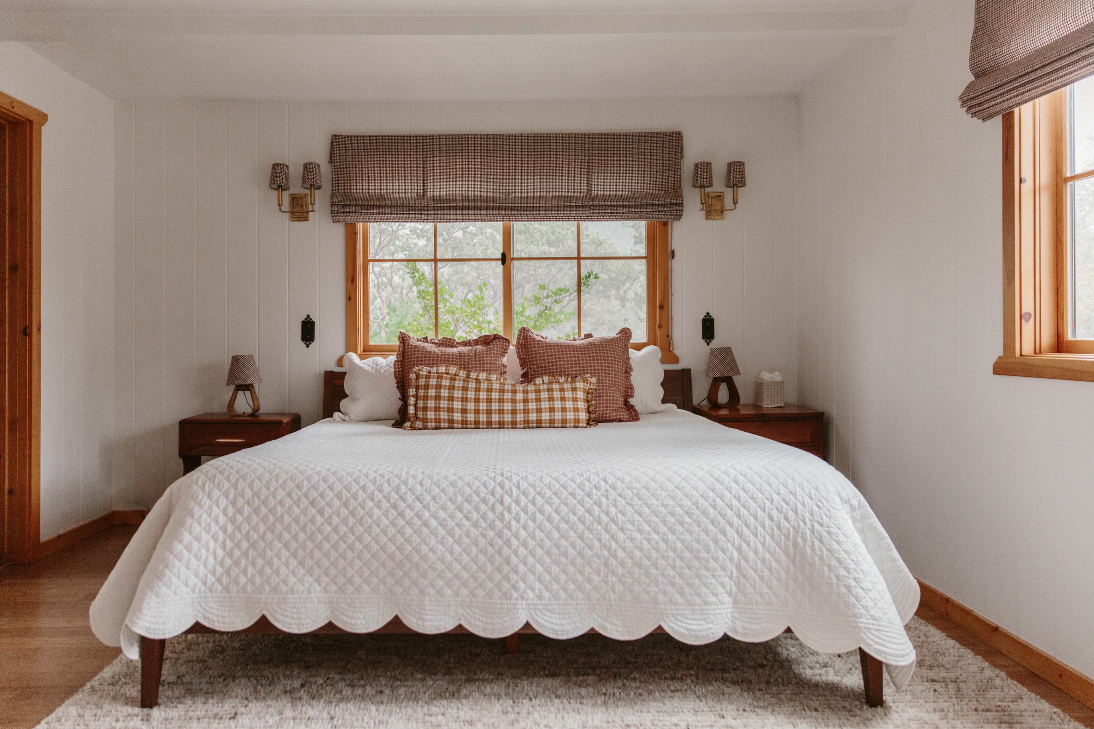 A neatly made bed with scalloped edges and decorative pillows is centered in a bright, minimalist bedroom featuring wooden accents and windows.