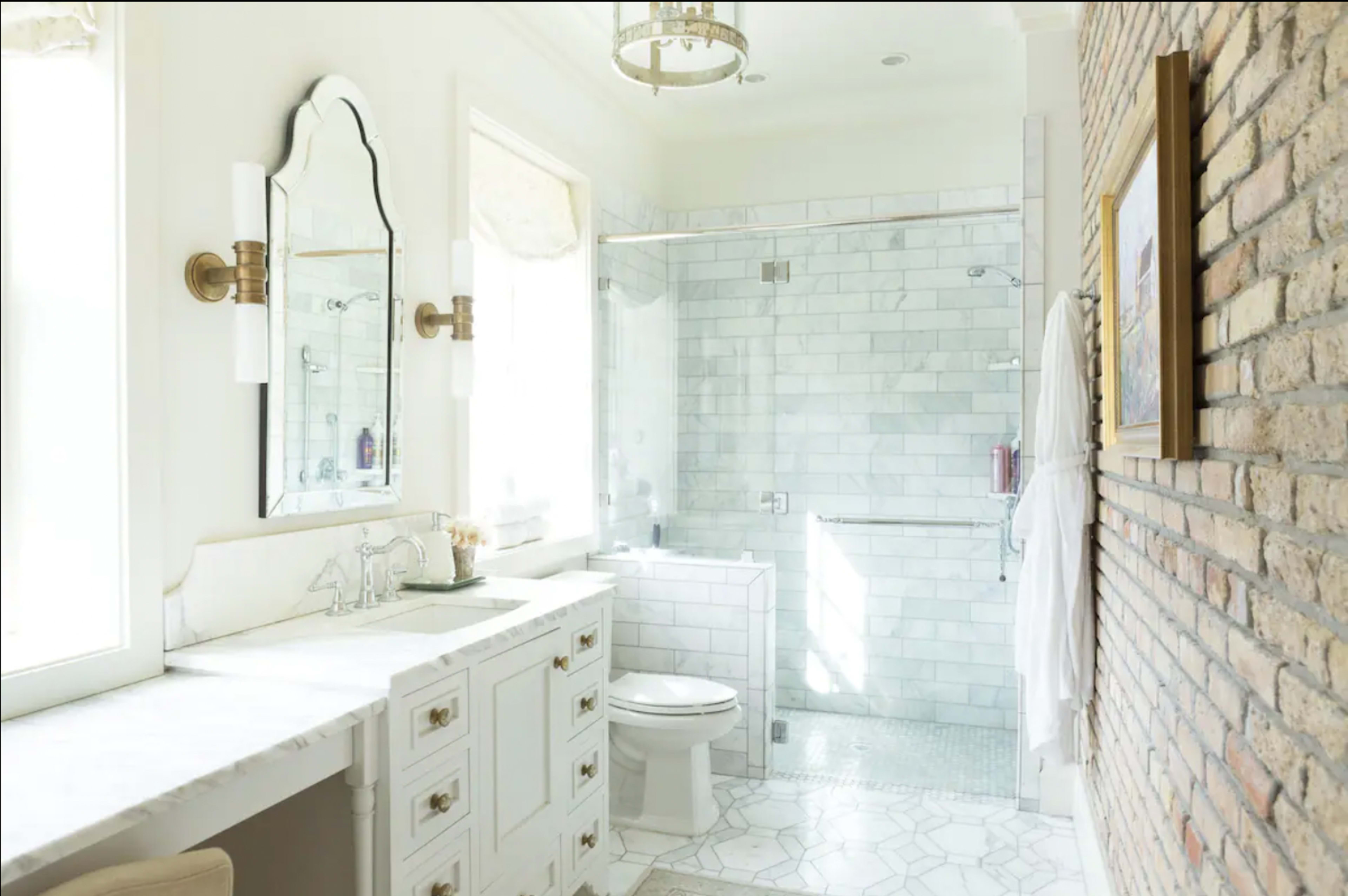 The bathroom features a marble-tiled shower area, a white vanity with drawers, and exposed brick accents on one wall.