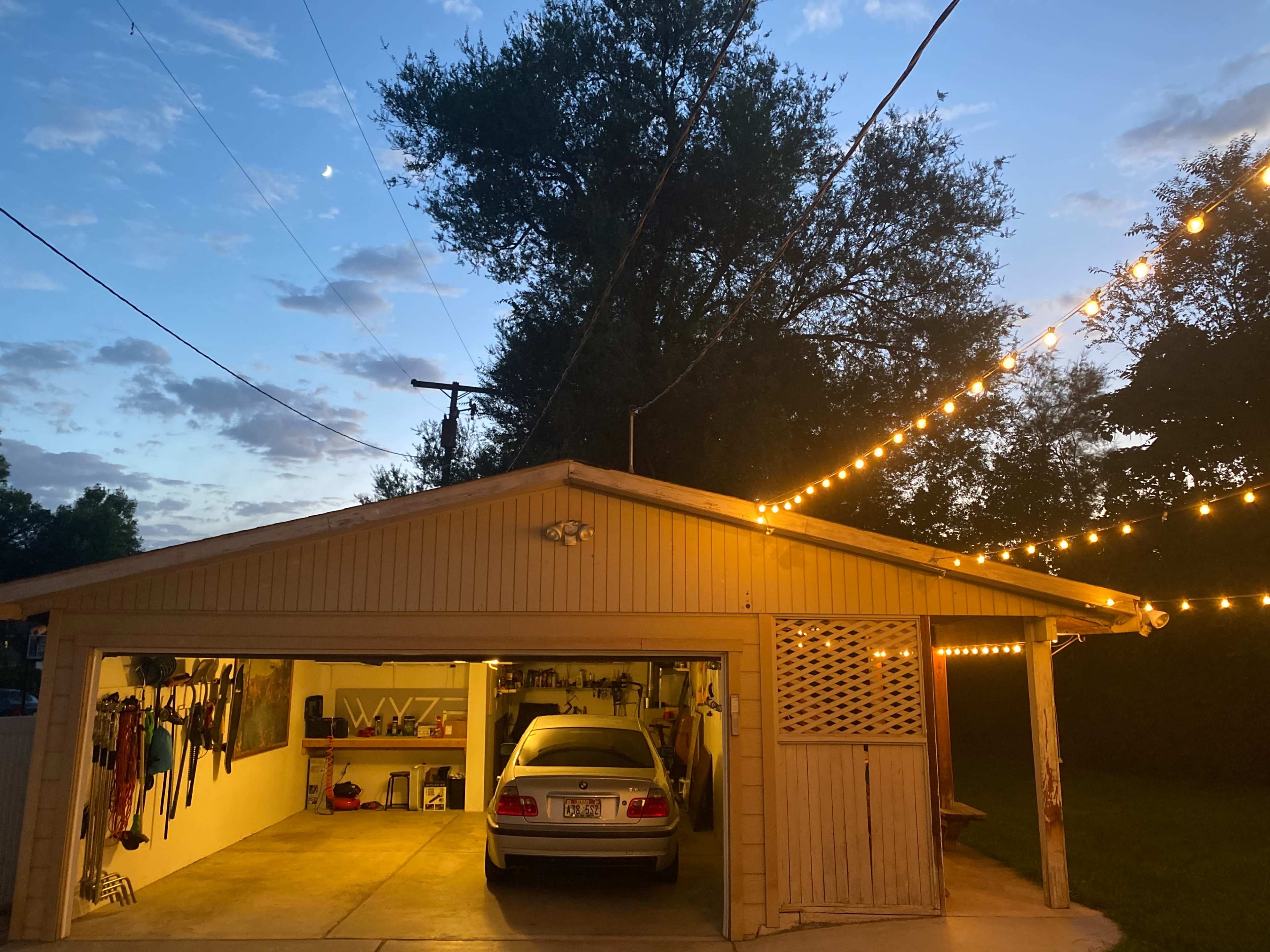 A garage with a car parked inside is illuminated by string lights in the evening, under a sky with scattered clouds and a visible moon.