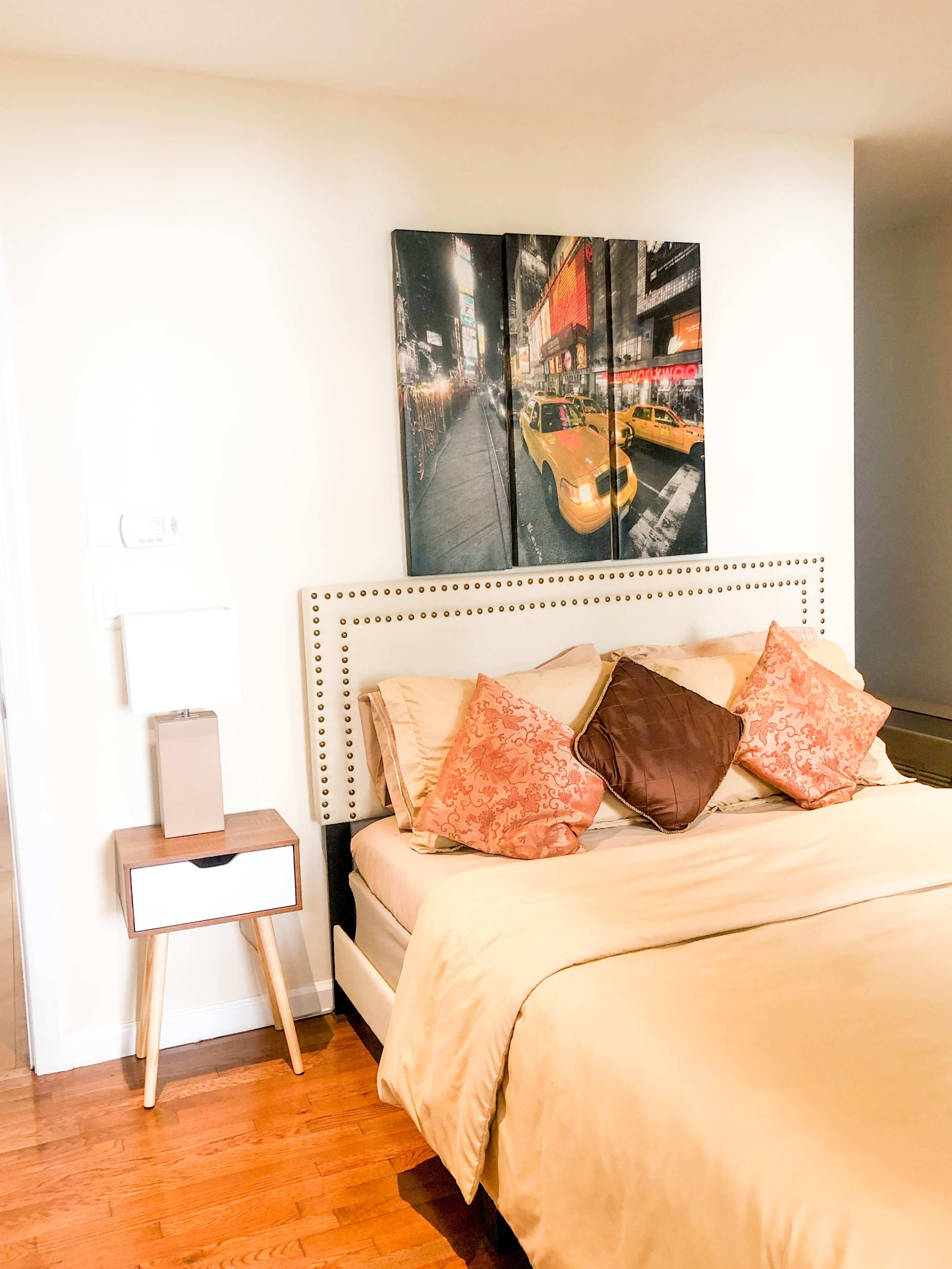 A bedroom featuring a bed with decorative pillows, a wooden nightstand with a lamp, and a large wall art piece depicting a city scene.